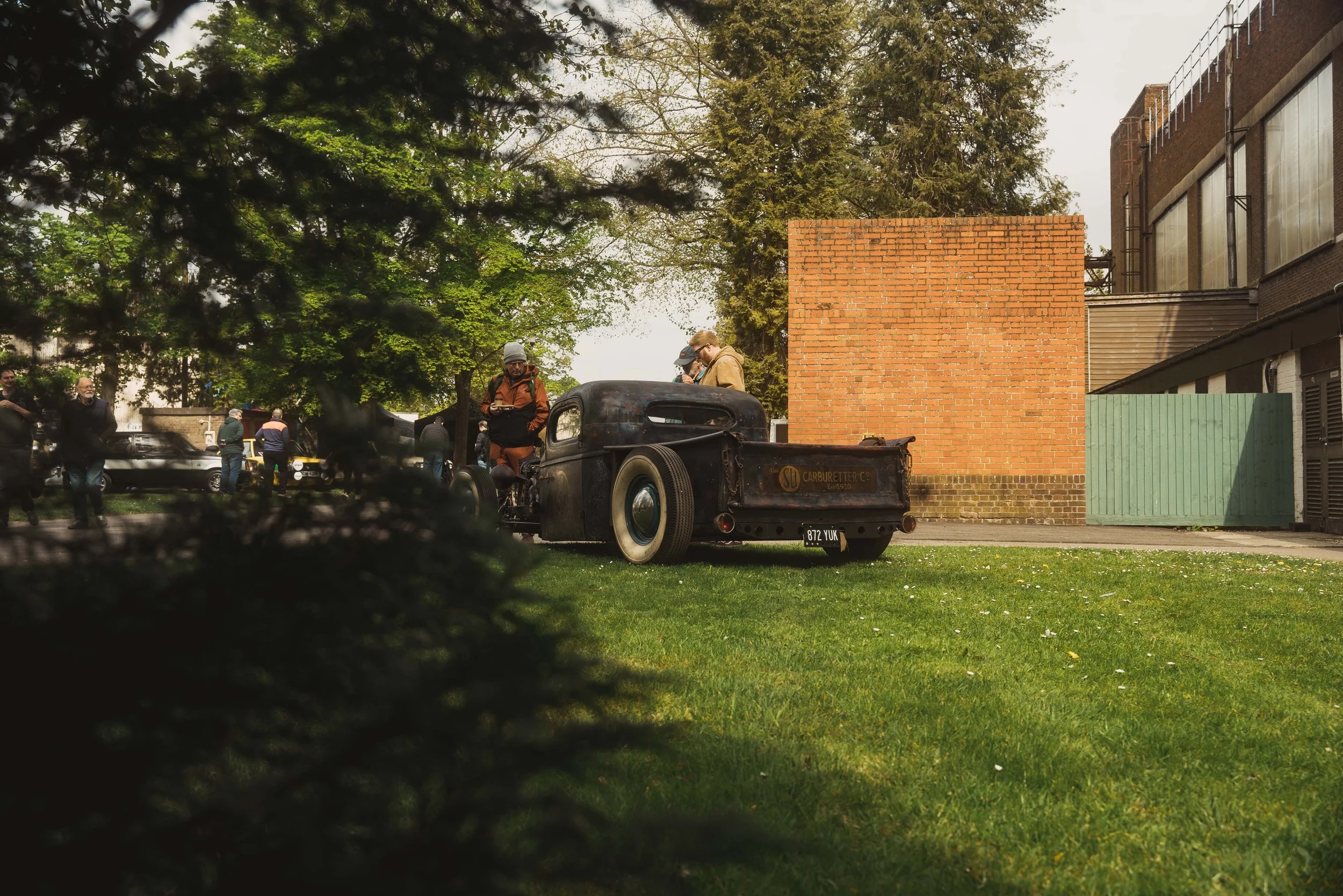 Vintage black car with rust and old signs, parked on a grassy area behind a tree, with people standing around and looking at it, in an outdoor setting with trees, brick, and metal buildings.