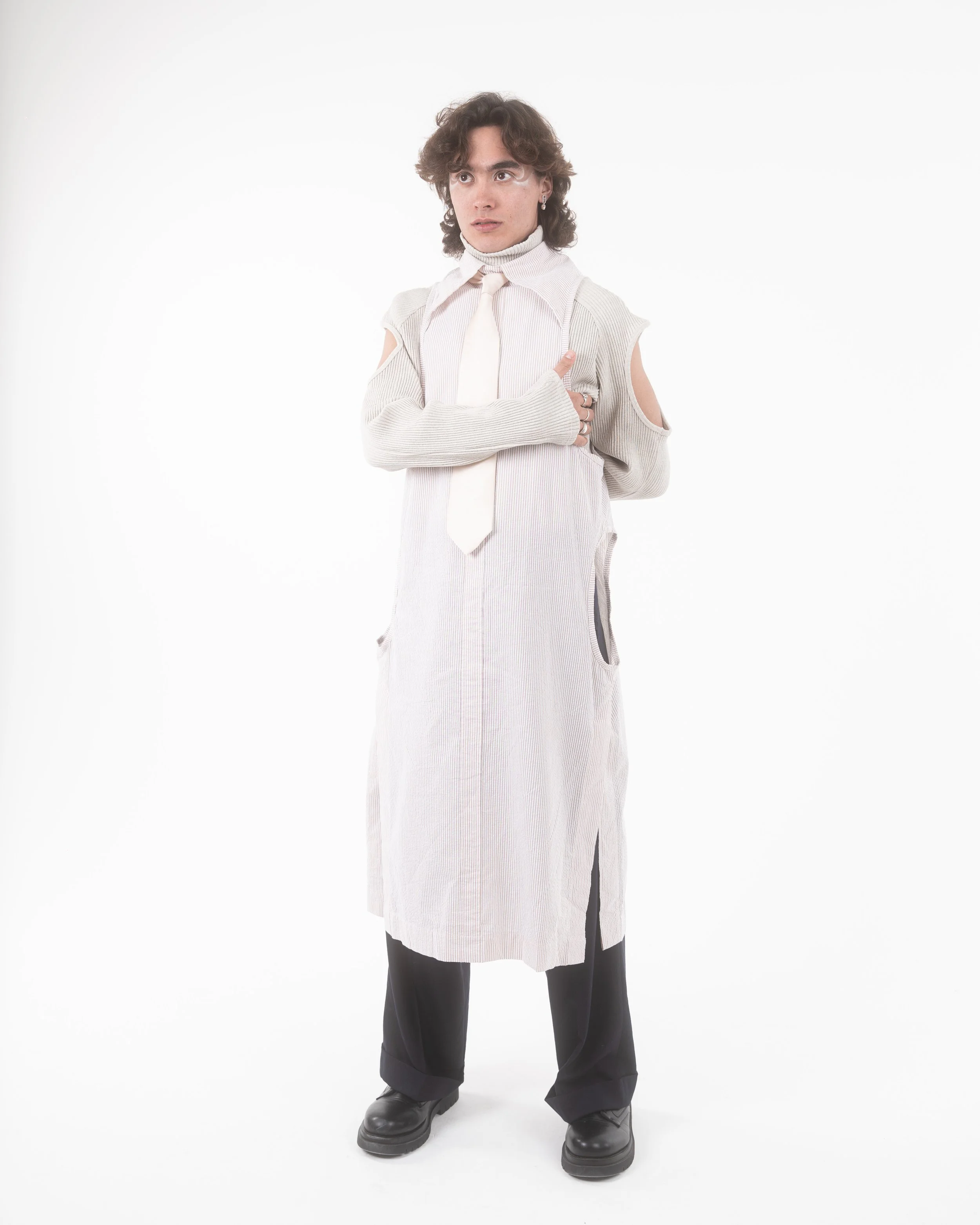 Person with curly hair wearing a gray open-shoulder sweater, white tie, and black shoes, standing against a white background.