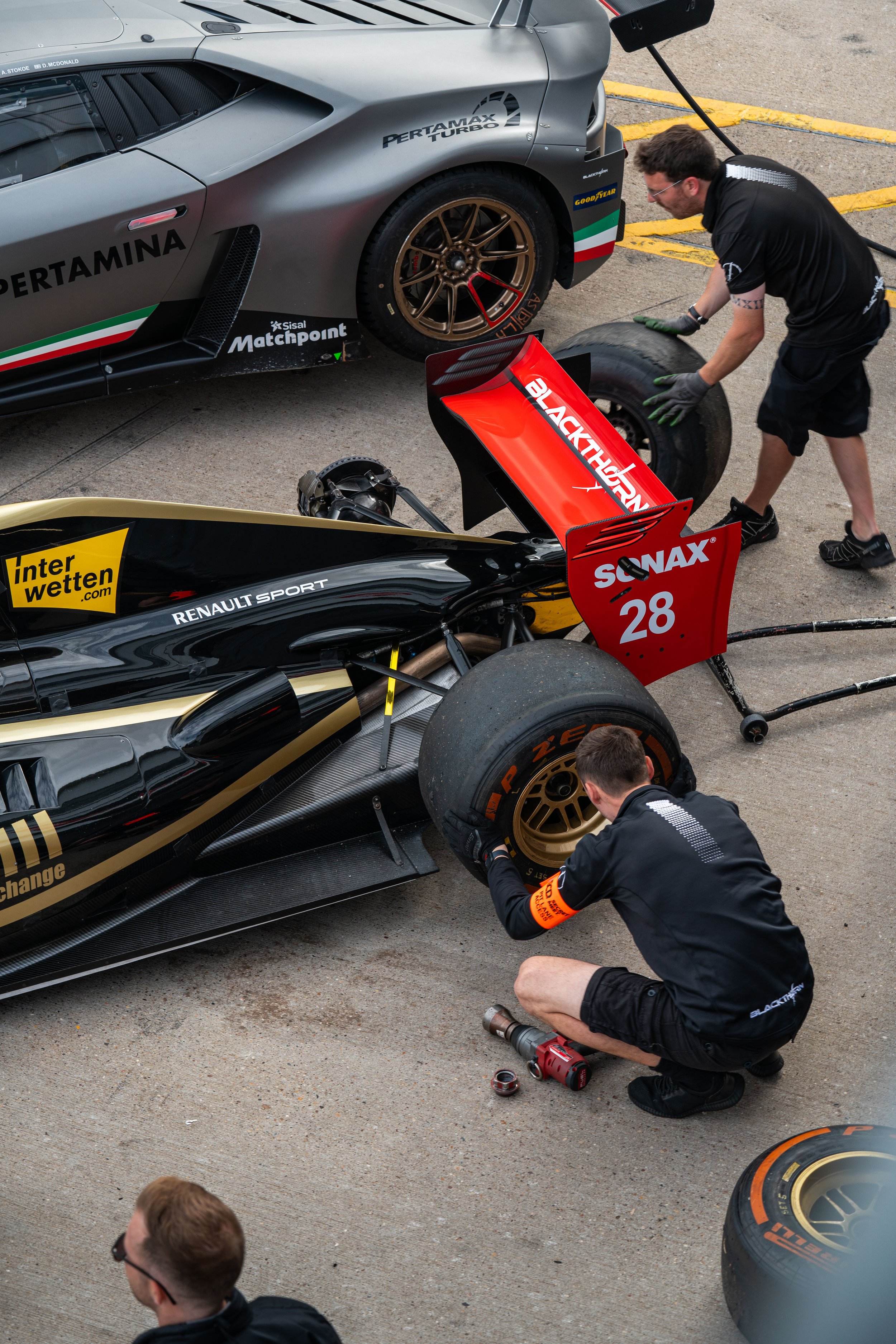 Race car team members working on a black and gold Renault Formula race car in the pit lane, changing tires and inspecting the car.