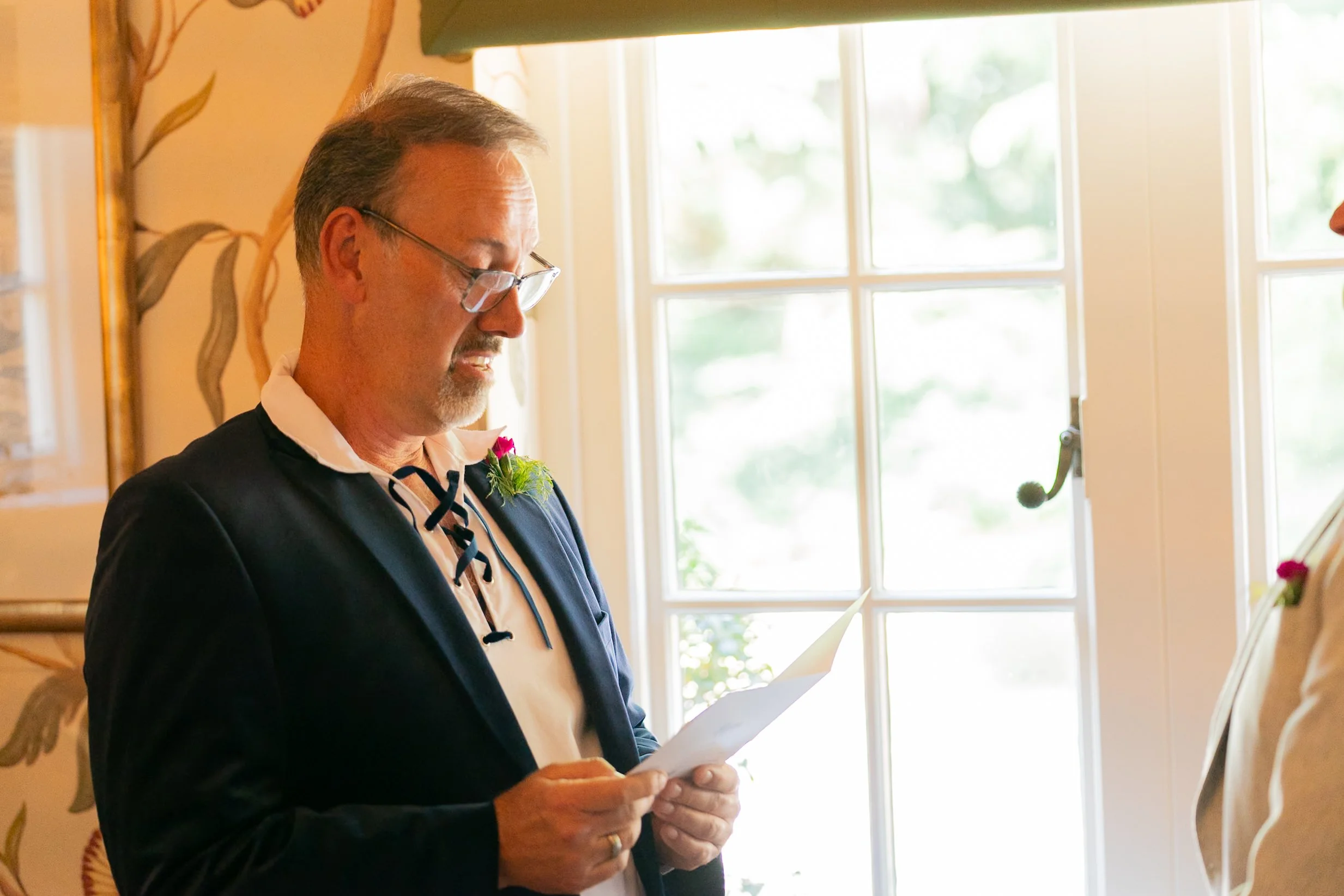 A man dressed in traditional attire, reading from a paper during a ceremony, with a window in the background and a boutonniere on his chest.