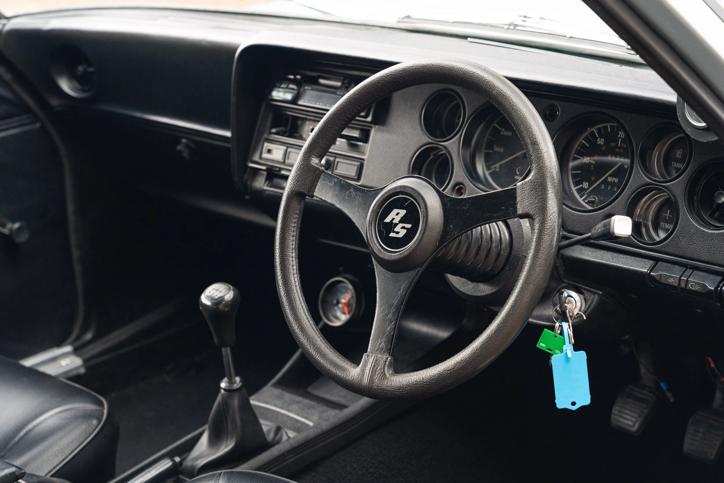 Interior of a car with a black dashboard, steering wheel with 'RS' logo, manual gear shift, and instrument cluster showing speed, RPM, and other gauges.