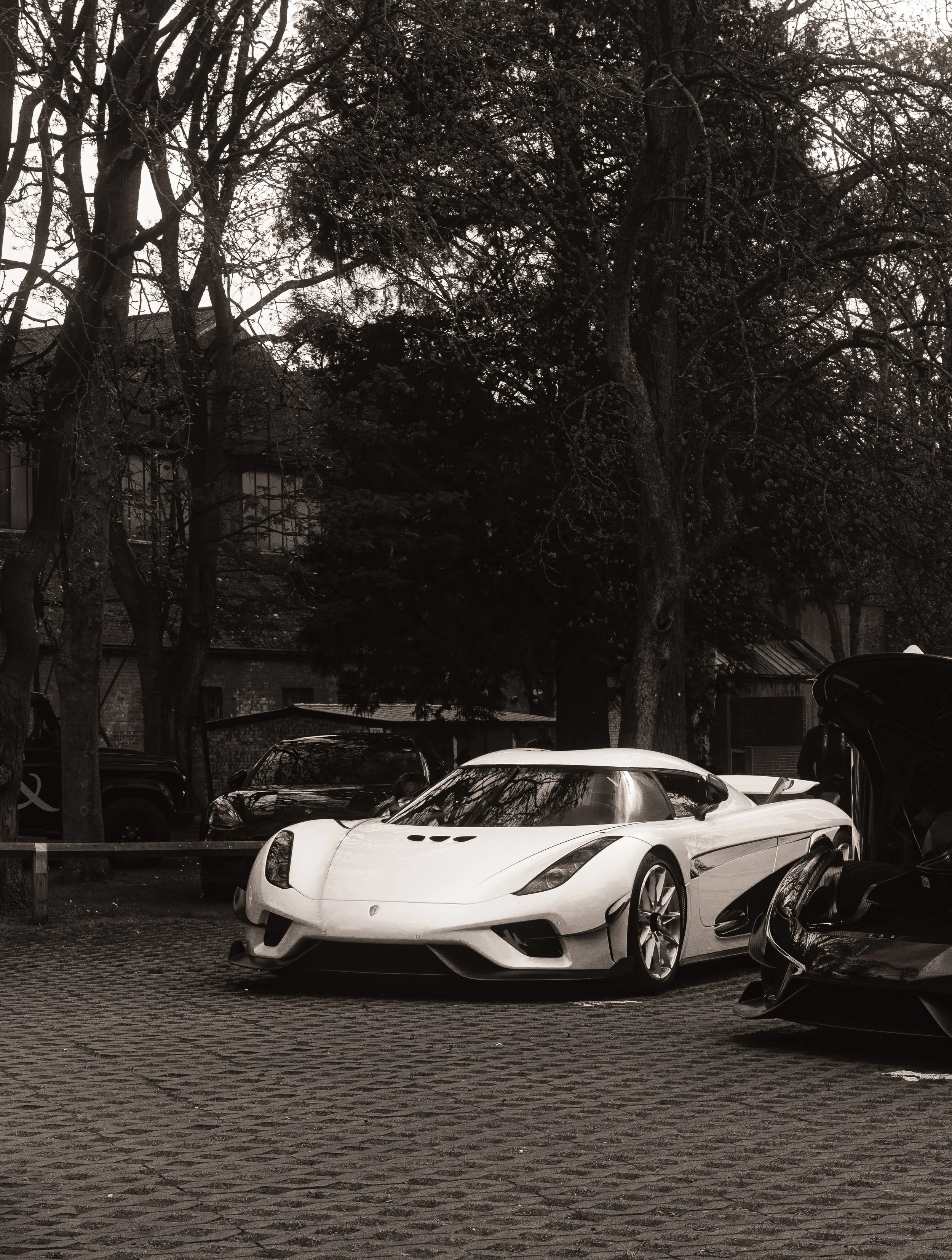 A white luxury sports car parked on a cobblestone street, with other cars and trees in the background, in a black and white photograph.