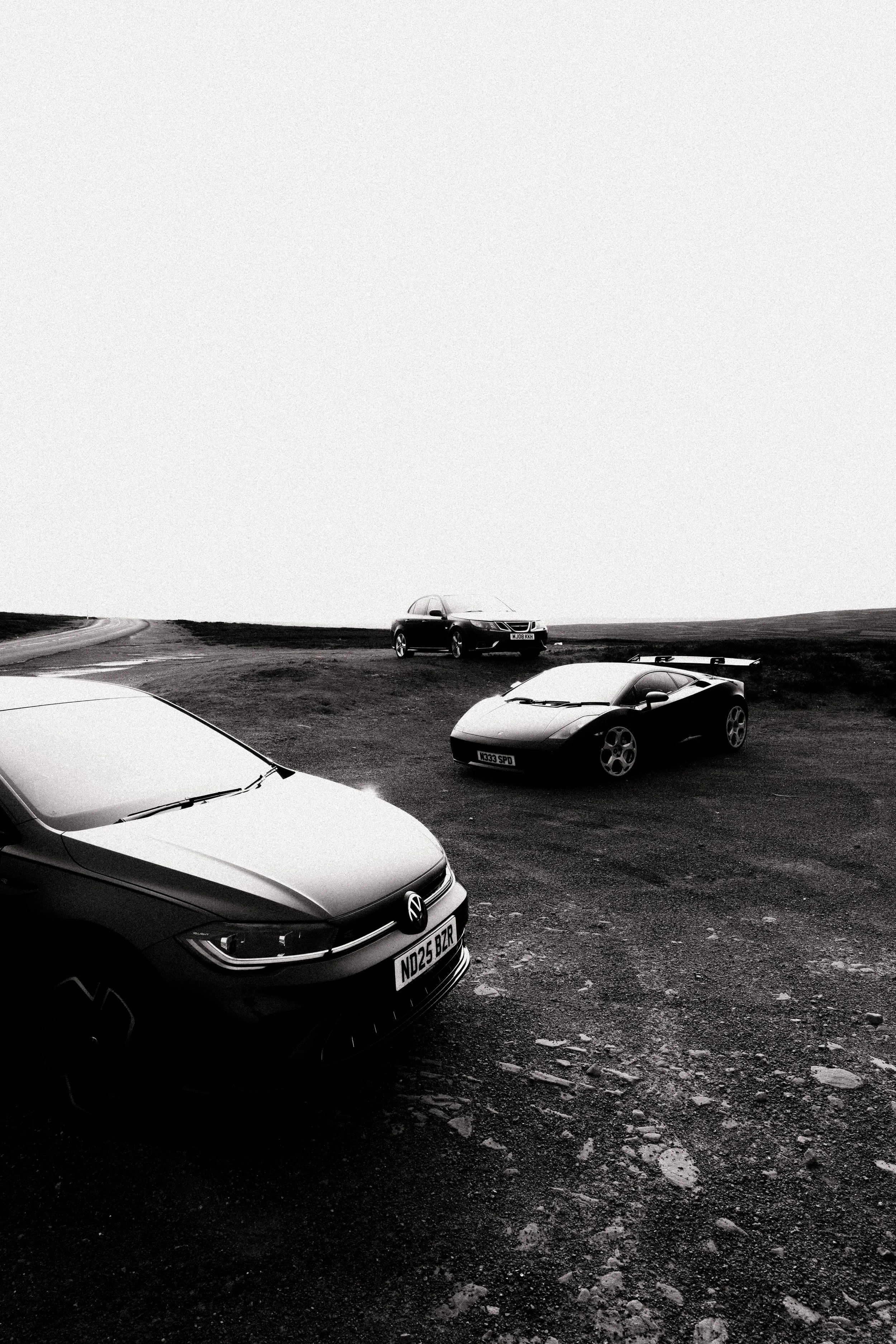 Three cars parked on a rough grassy hilltop in black and white, with open sky and distant landscape