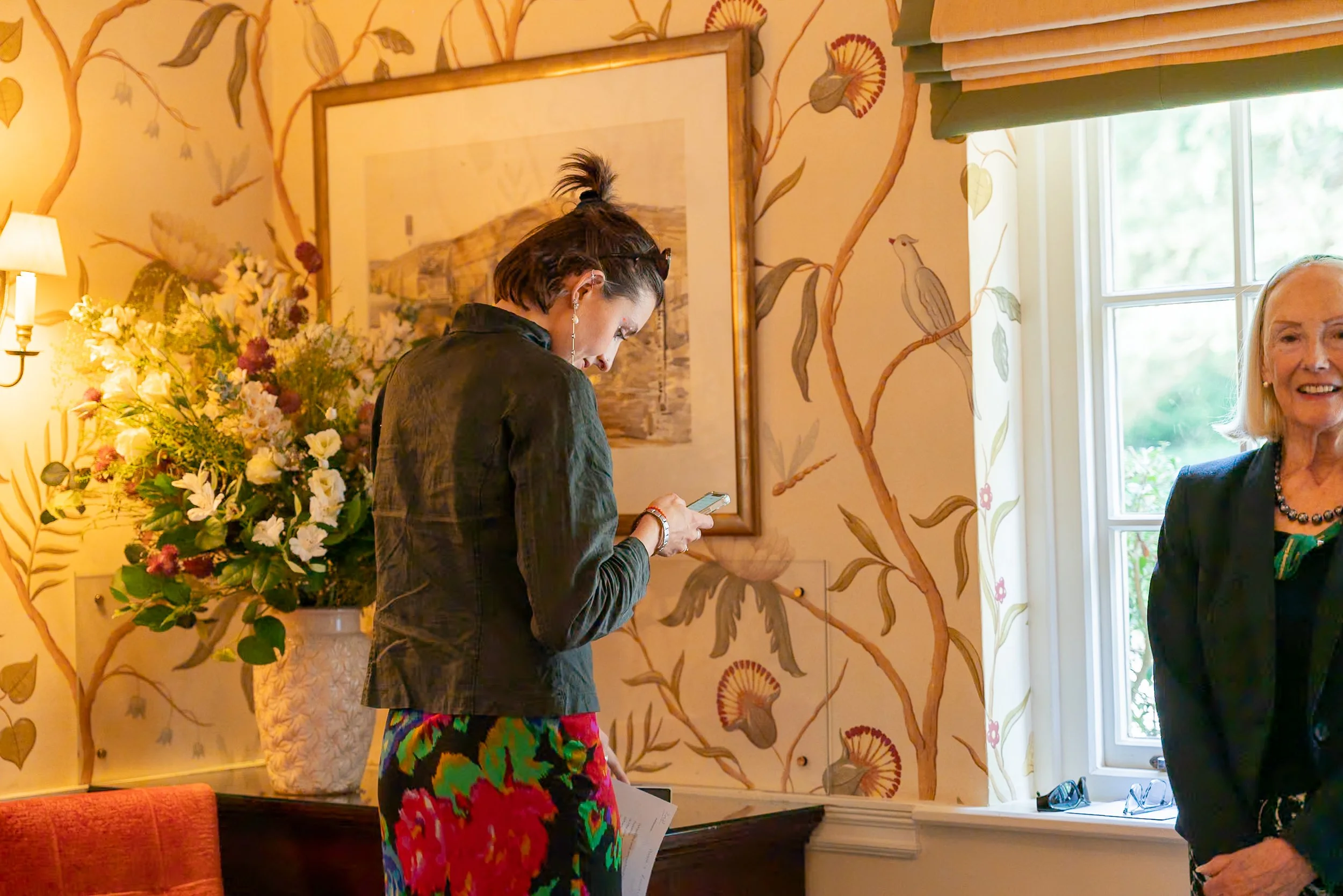 Two women, one with short dark hair looking at her phone and the other with shoulder-length blonde hair smiling, standing in a decorated room with floral wallpaper, a large floral arrangement, and a window with blinds.