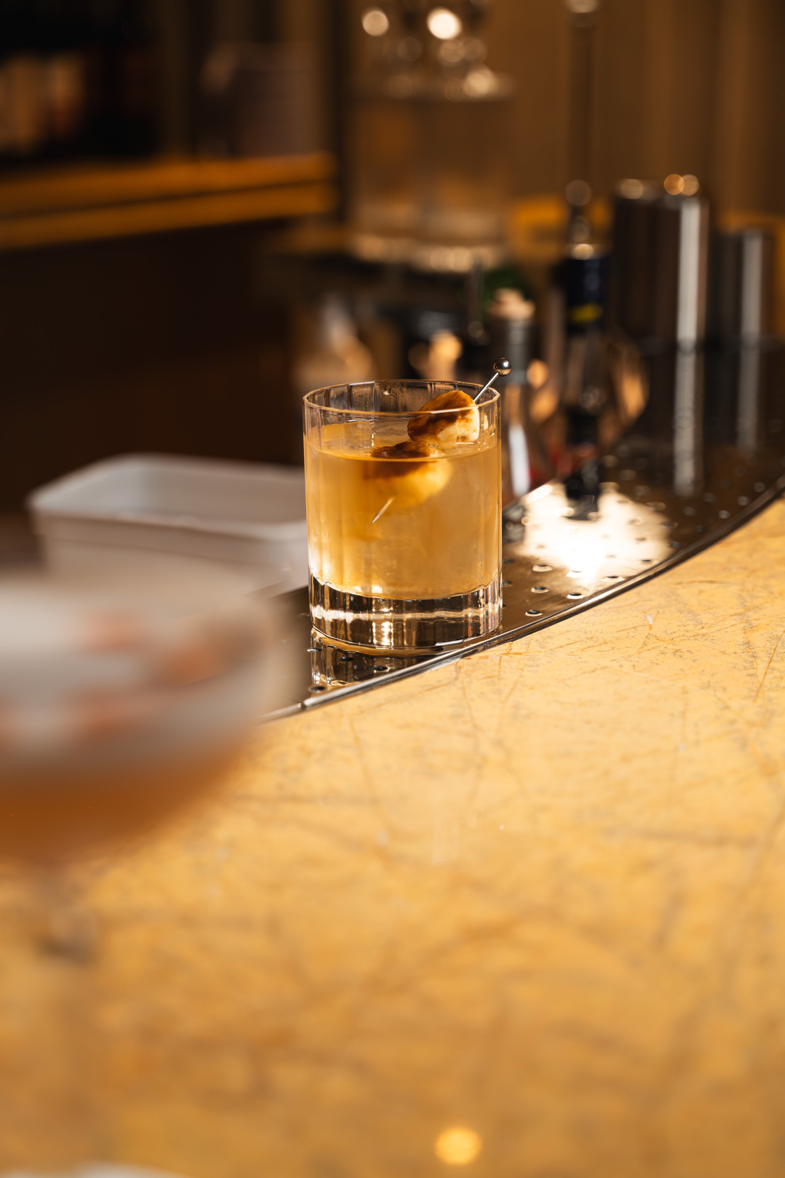 A close-up of a glass of whiskey with a cherry garnish on a bar counter, out of focus background of bar tools and shelves.