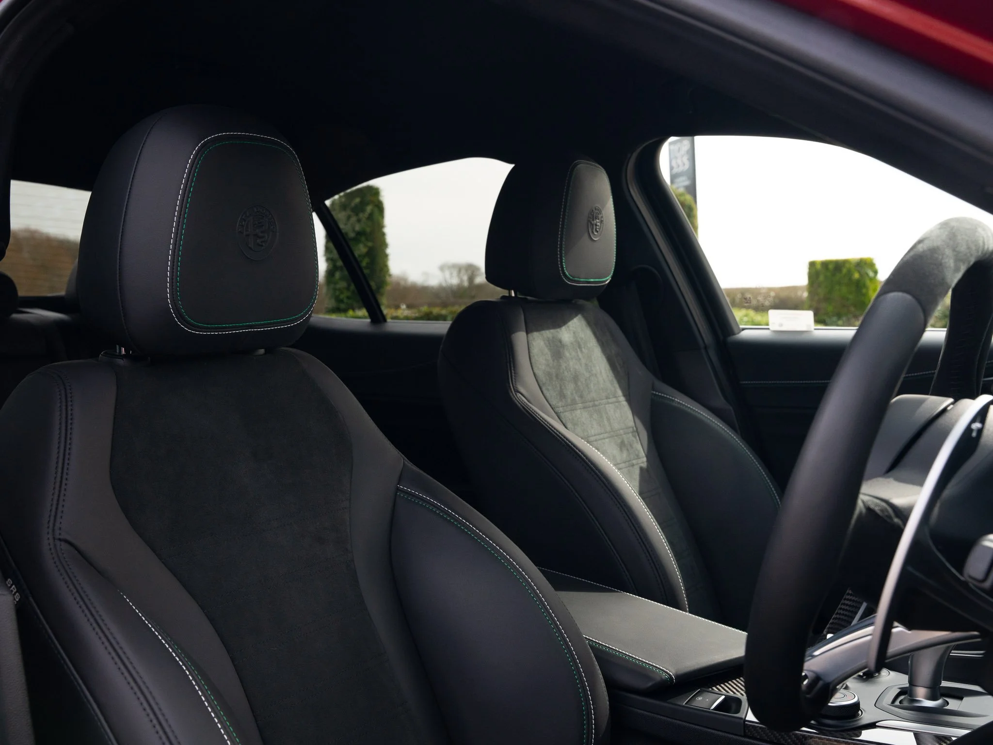 Inside view of a car with black leather front seats, dashboard, and steering wheel, with a view of greenery outside