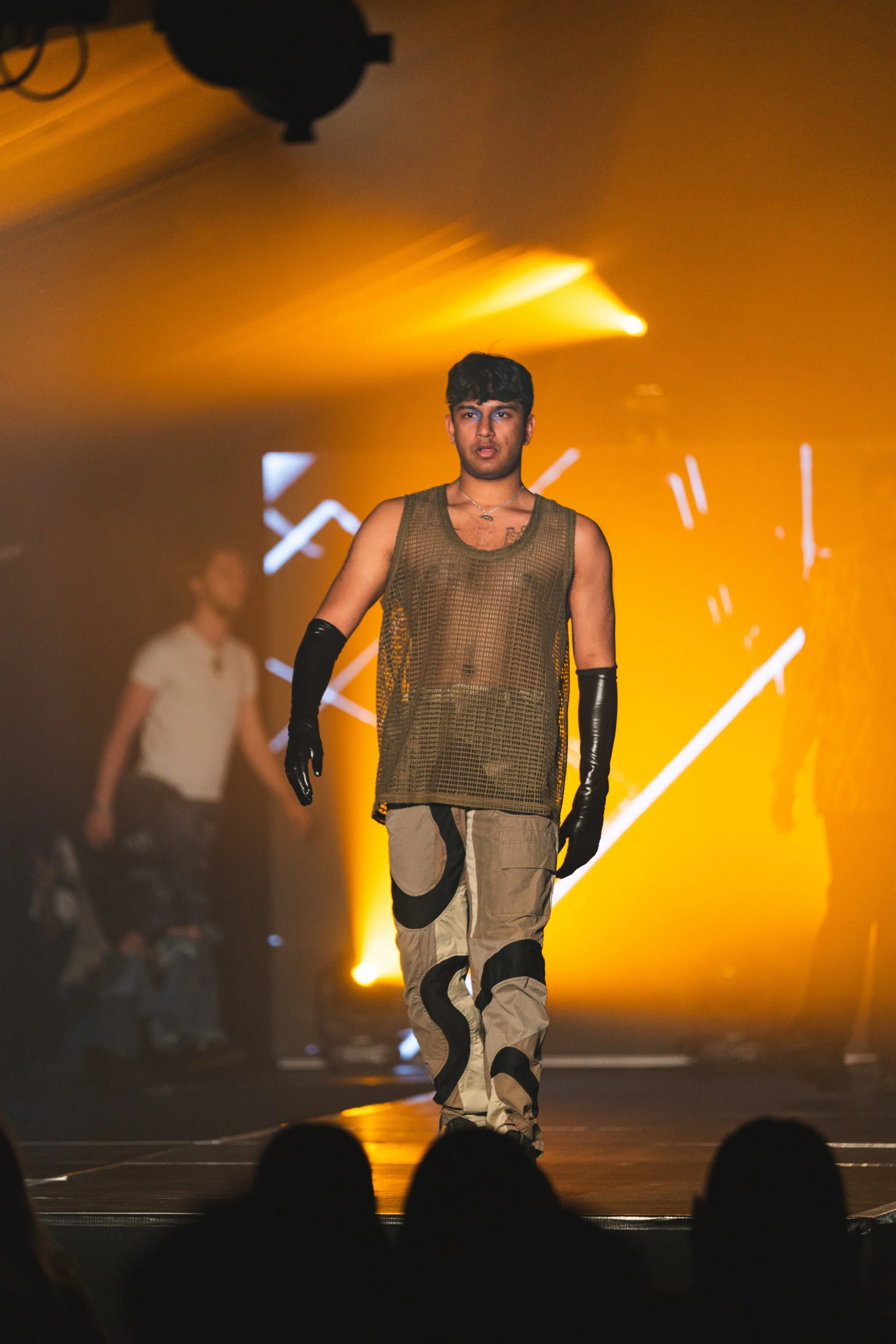Male model walking on runway during fashion show, wearing a mesh/tank top and stylish pants with black accents, black gloves, with vibrant yellow stage lighting and abstract background graphics.