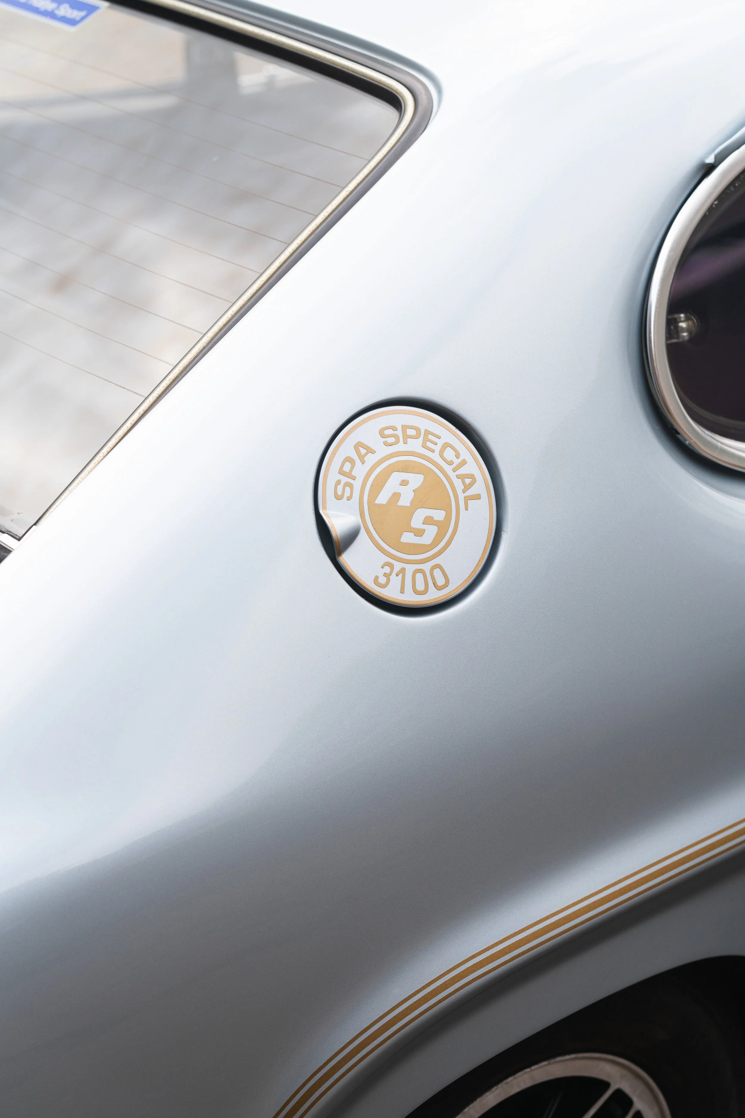 Close-up of a vintage silver car with a circular badge reading "Spa Special RS 3100" near the rear window, with a rounded rear tail light partially visible.