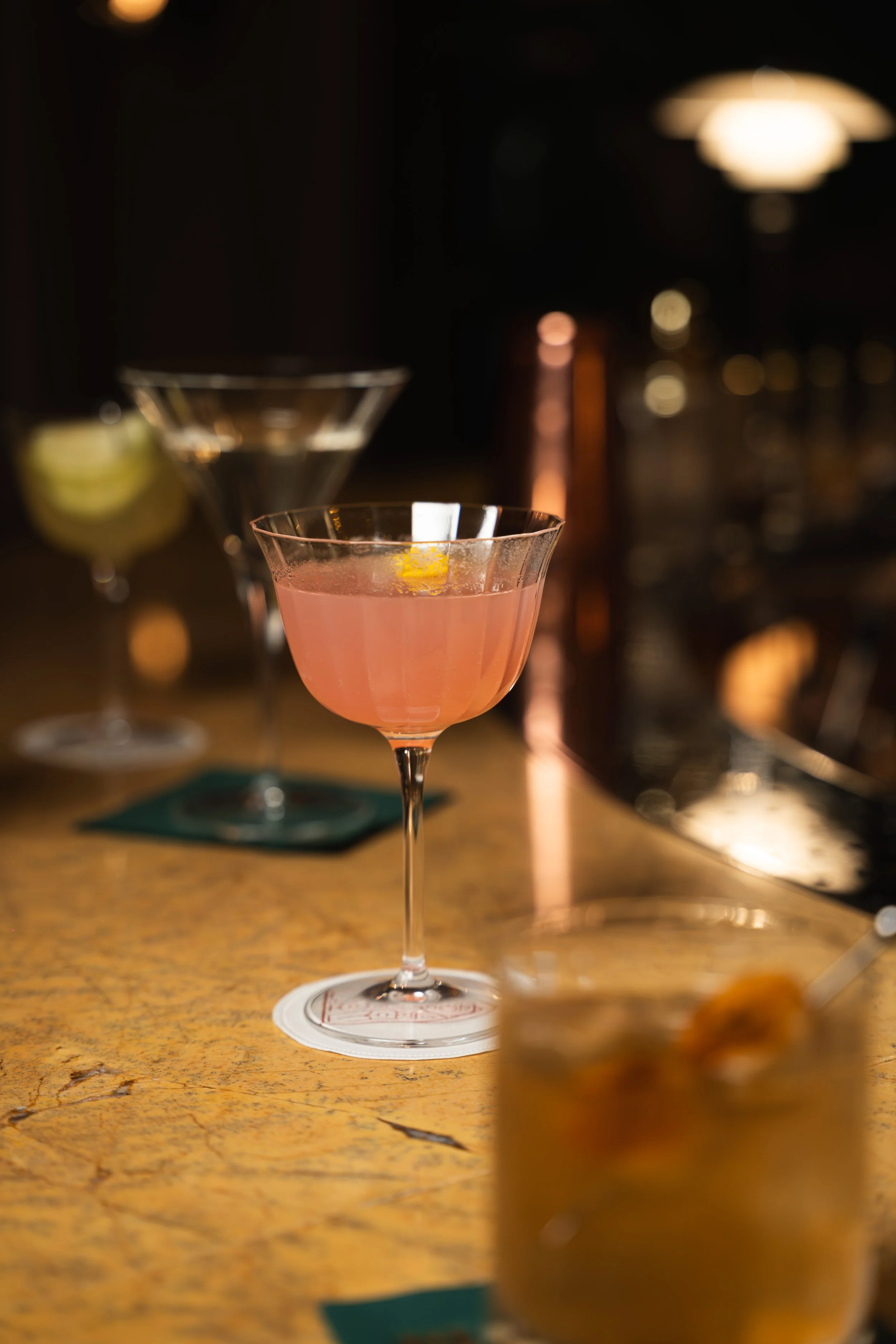 A pink cocktail with a lemon twist garnish in a stemmed glass on a bar counter.