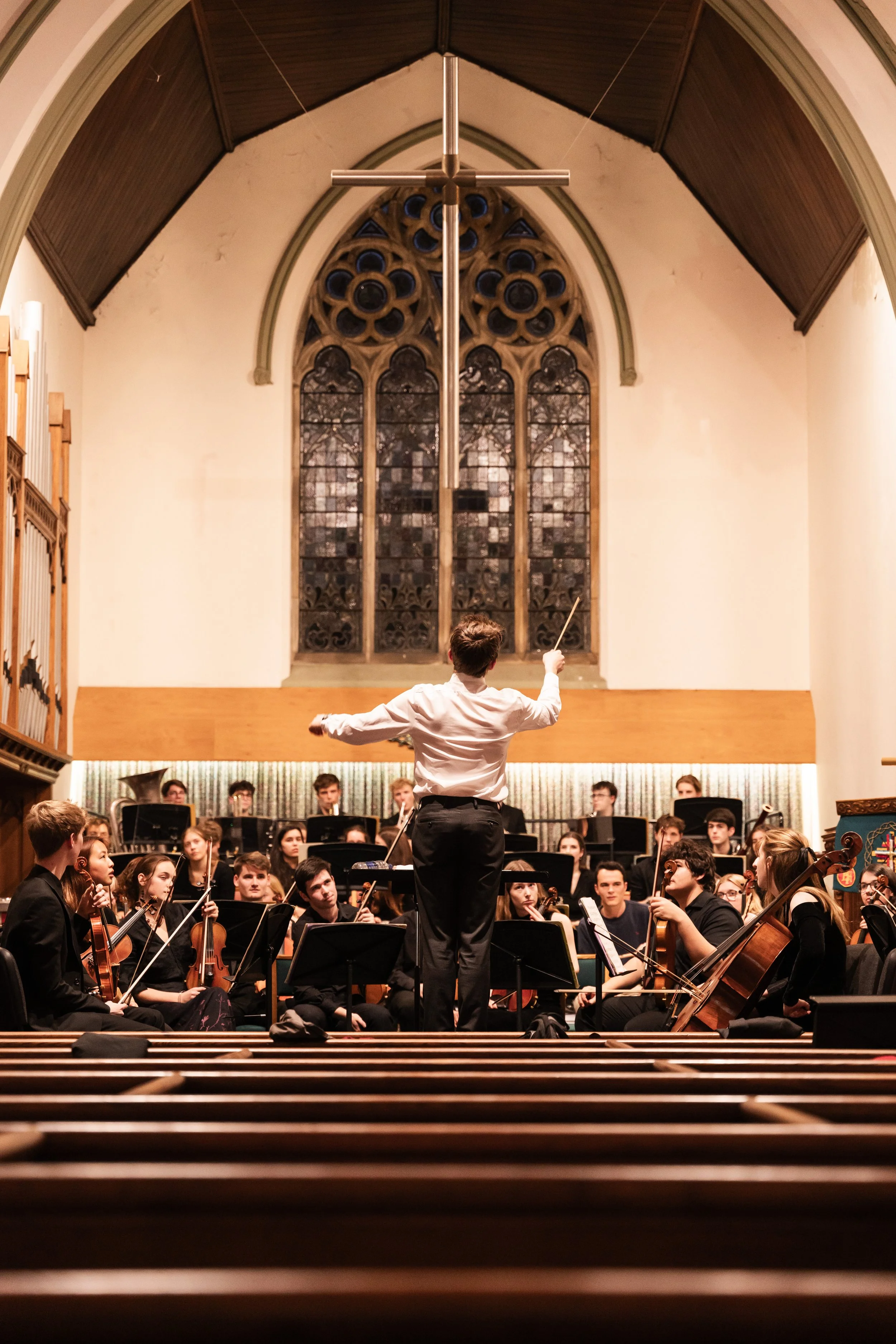 Orchestra conductor leading an orchestra during a performance in a church or concert hall with stained glass windows.
