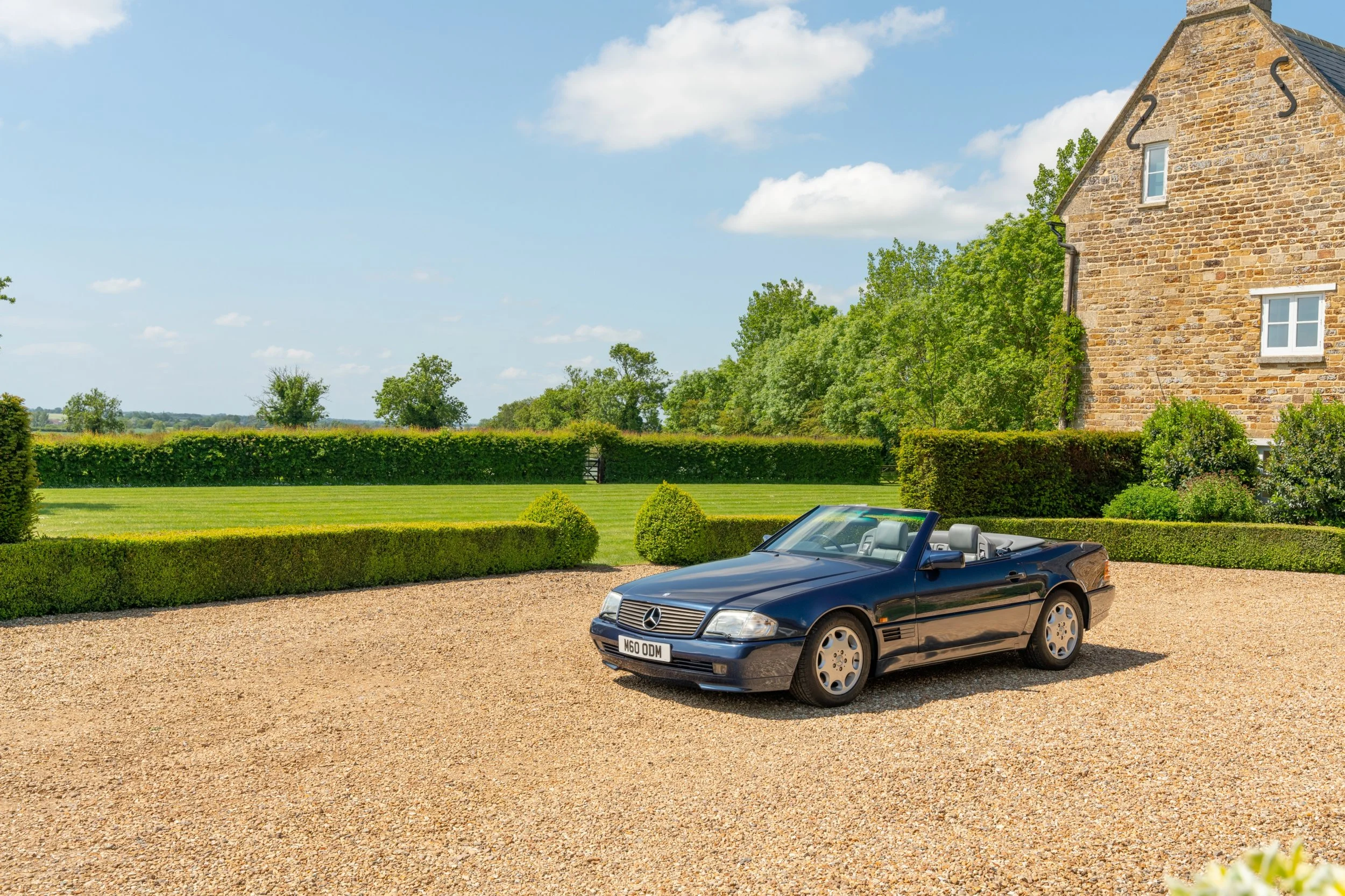 Black Mercedes-Benz convertible parked on gravel driveway in front of a stone house and manicured lawn, with trees and blue sky in the background.