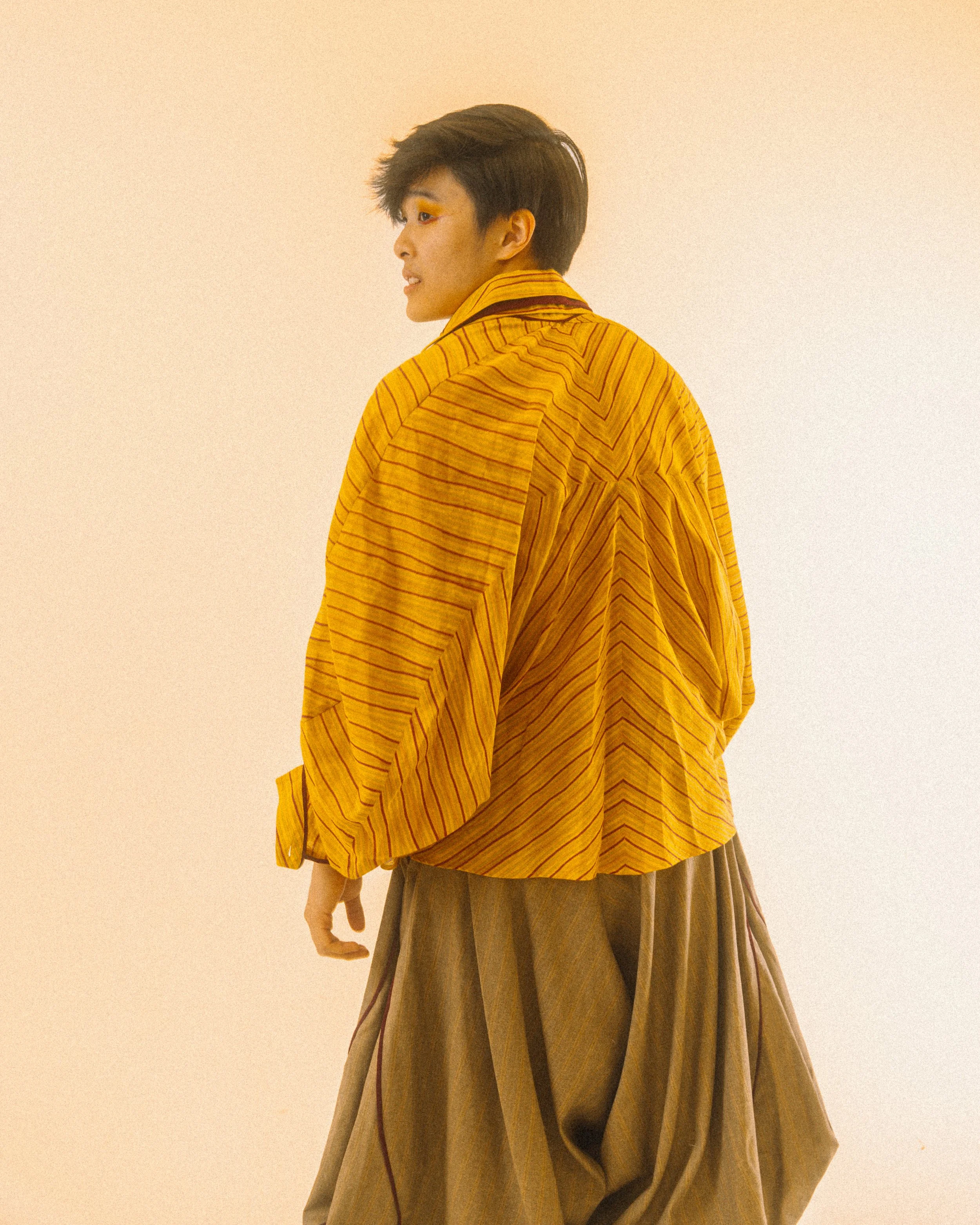 A person with short dark hair wearing a yellow and orange striped jacket and loose brown pants, standing against a plain beige background, looking to their left.