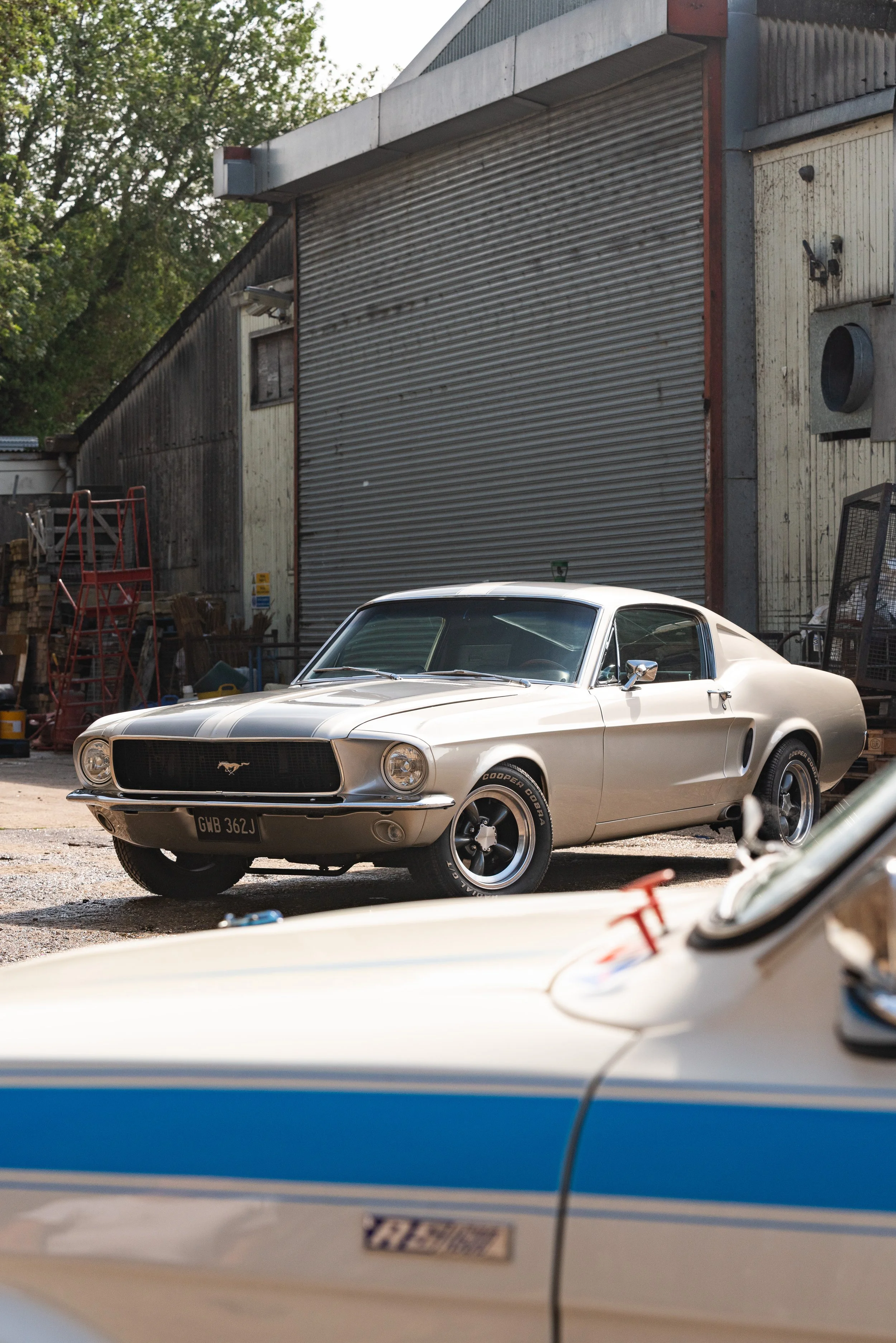 A classic silver Ford Mustang parked outside a rustic building with a closed metal roll-up door. There is another vintage car partially visible in the foreground.
