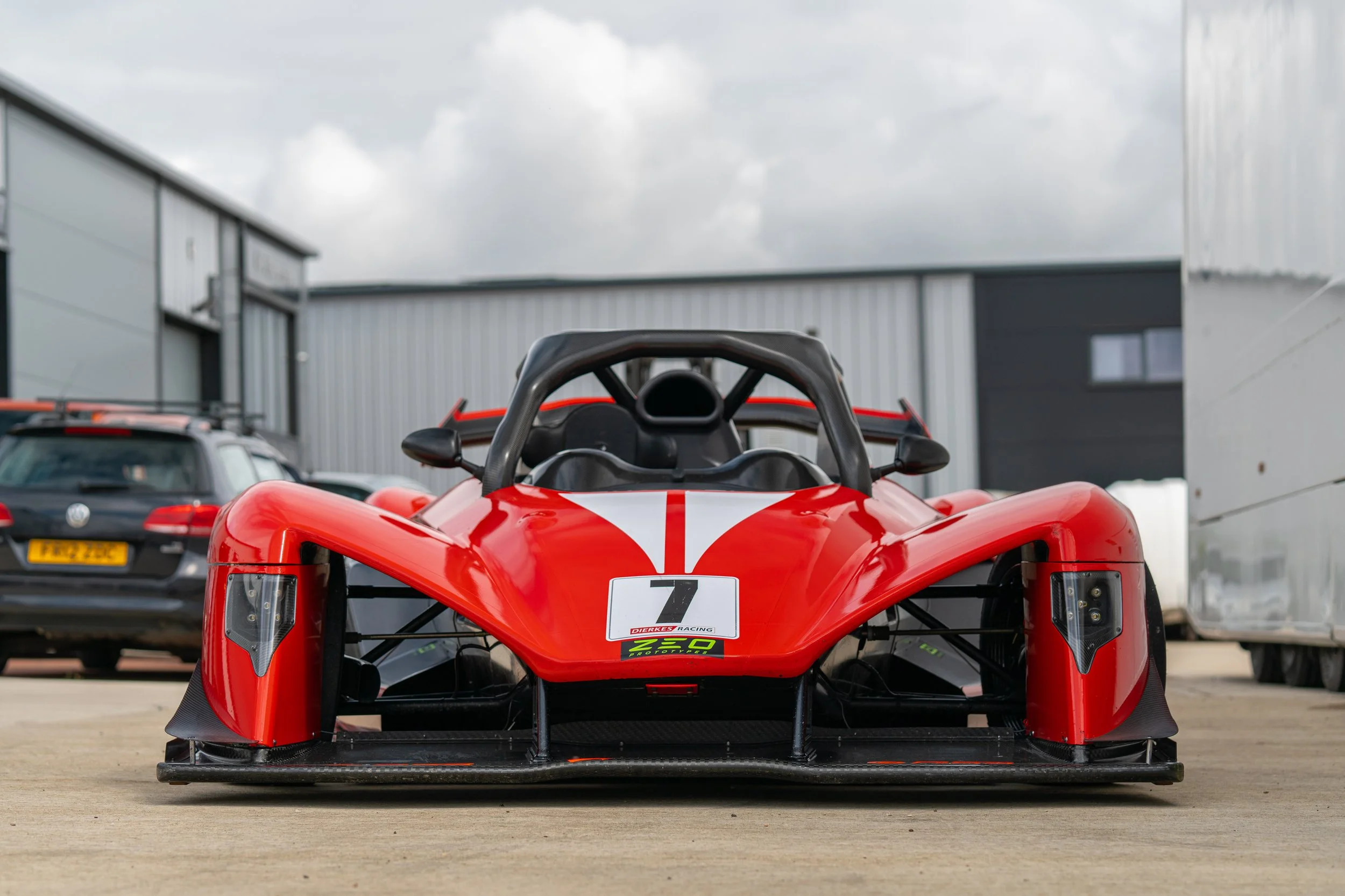 Red race car with black accents, number 7, parked outside garage with cars and industrial buildings in background.