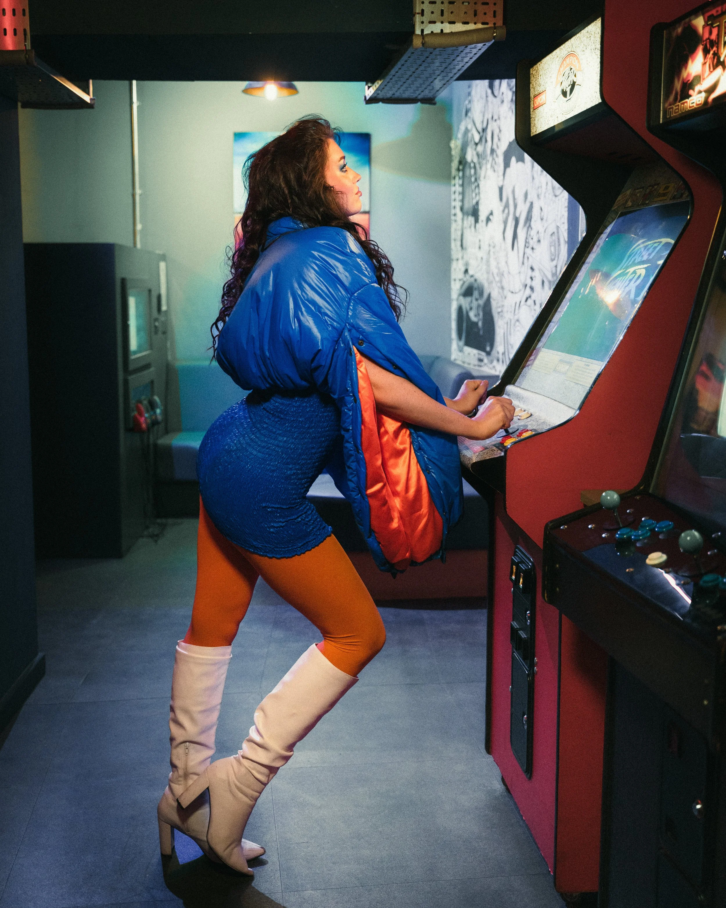 A woman playing a retro arcade game in a dimly lit arcade, wearing a blue jacket, a blue mini skirt, orange tights, and beige high-heeled boots.