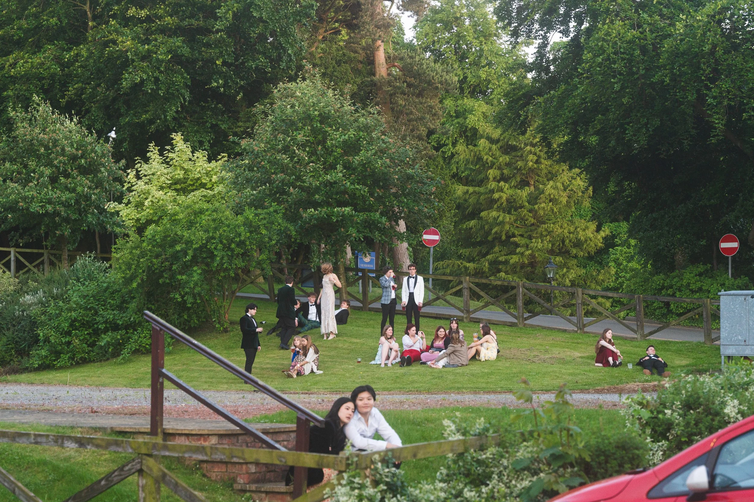 Group of people dressed formally, some seated on the grass, others standing and talking, in a park-like setting with trees and a wooden fence.