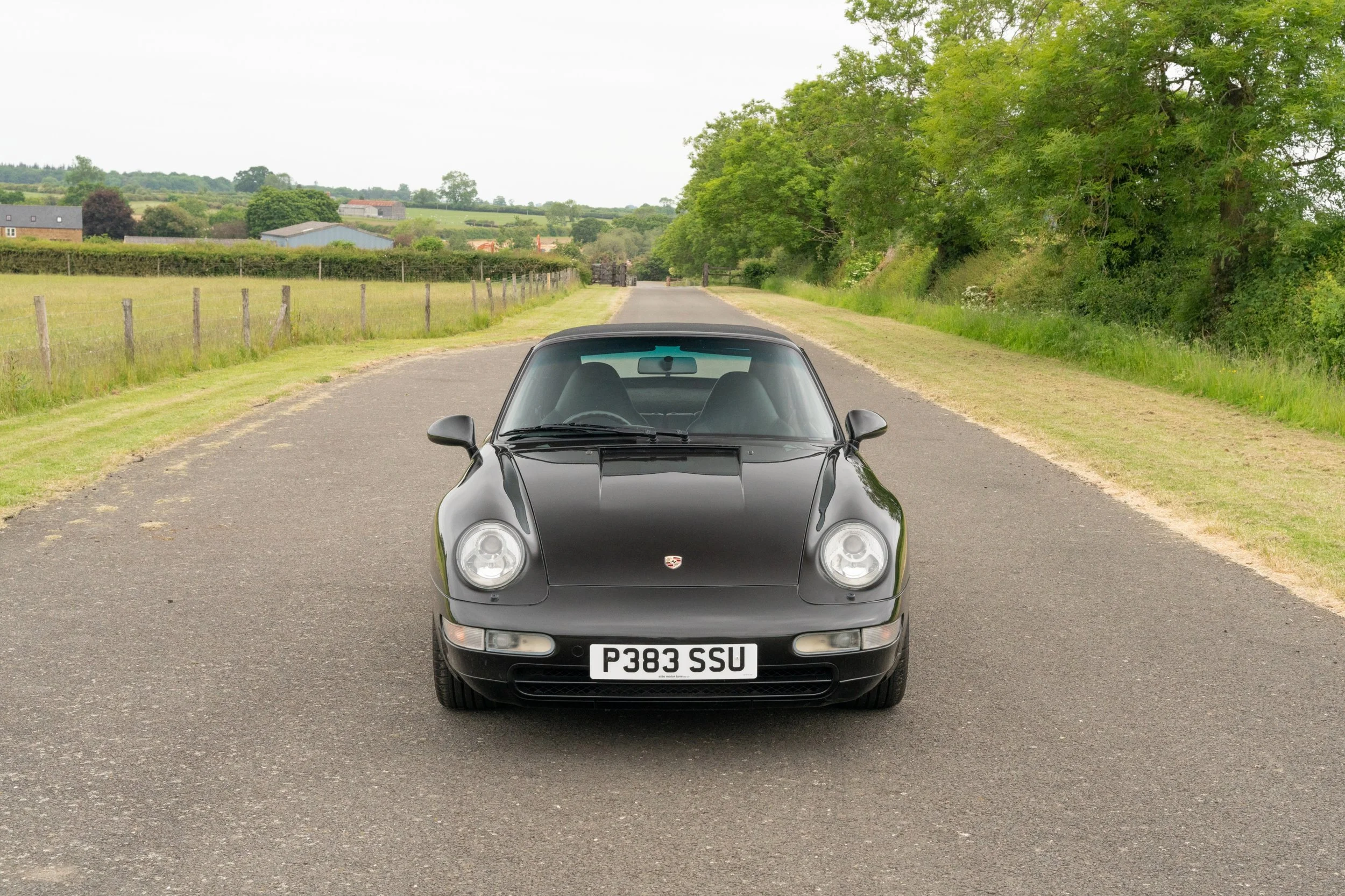 A black Porsche sports car parked on a rural road with grassy shoulders and trees on the right, and a fence with fields and houses in the background