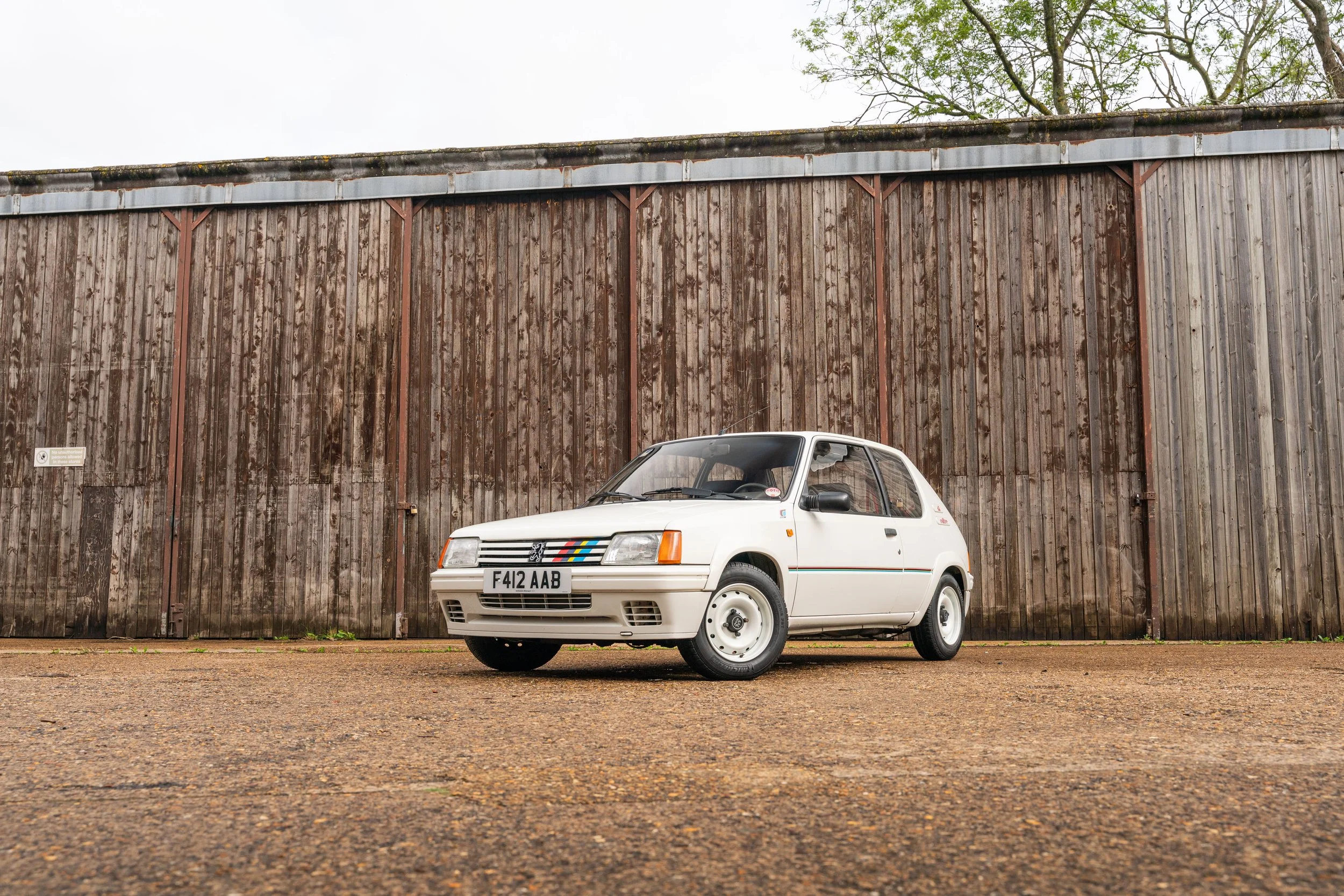 A white vintage hatchback car parked on a gravel surface in front of a weathered wooden wall.