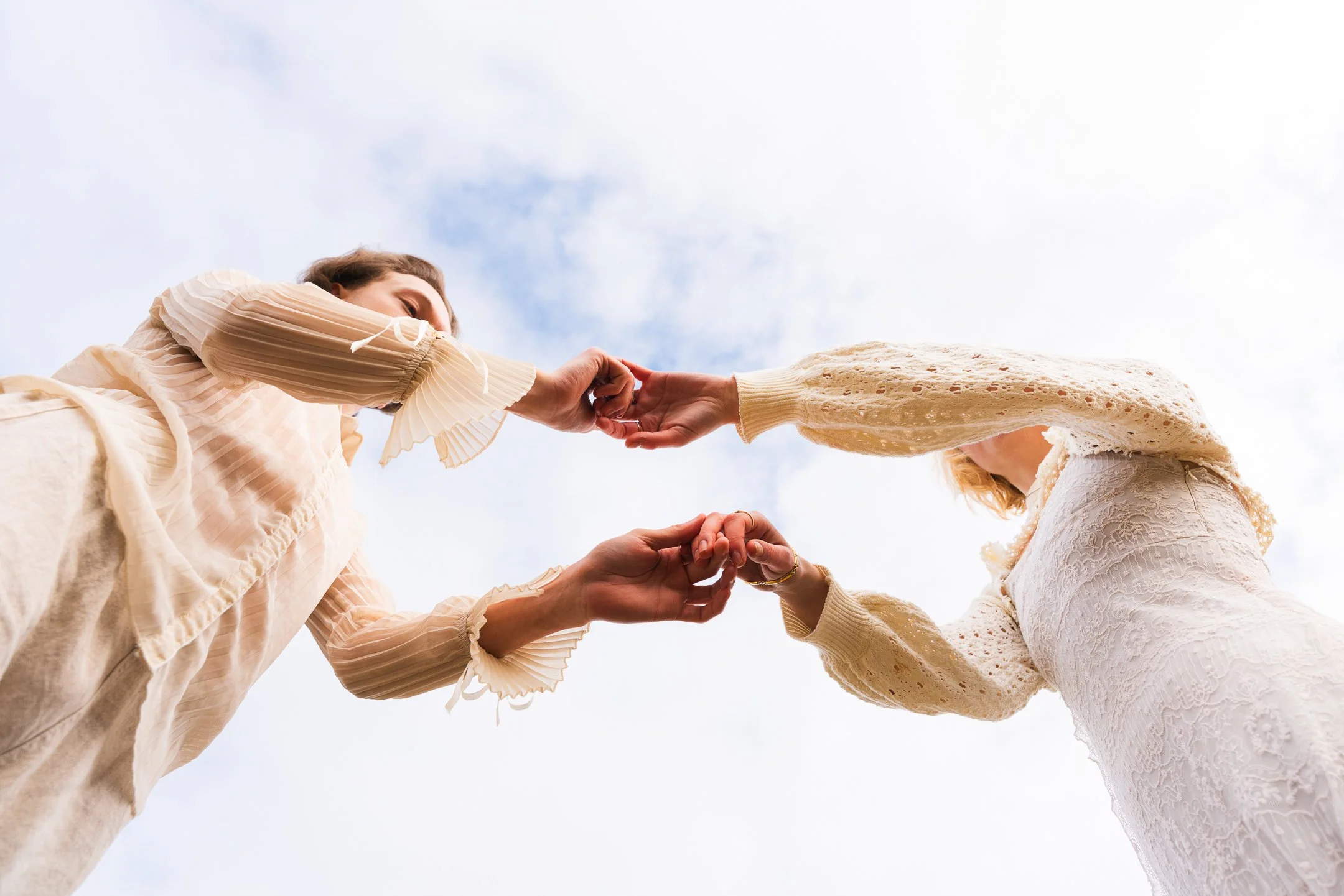 Two women in cream-colored clothing holding hands and touching fingers against a cloudy sky.