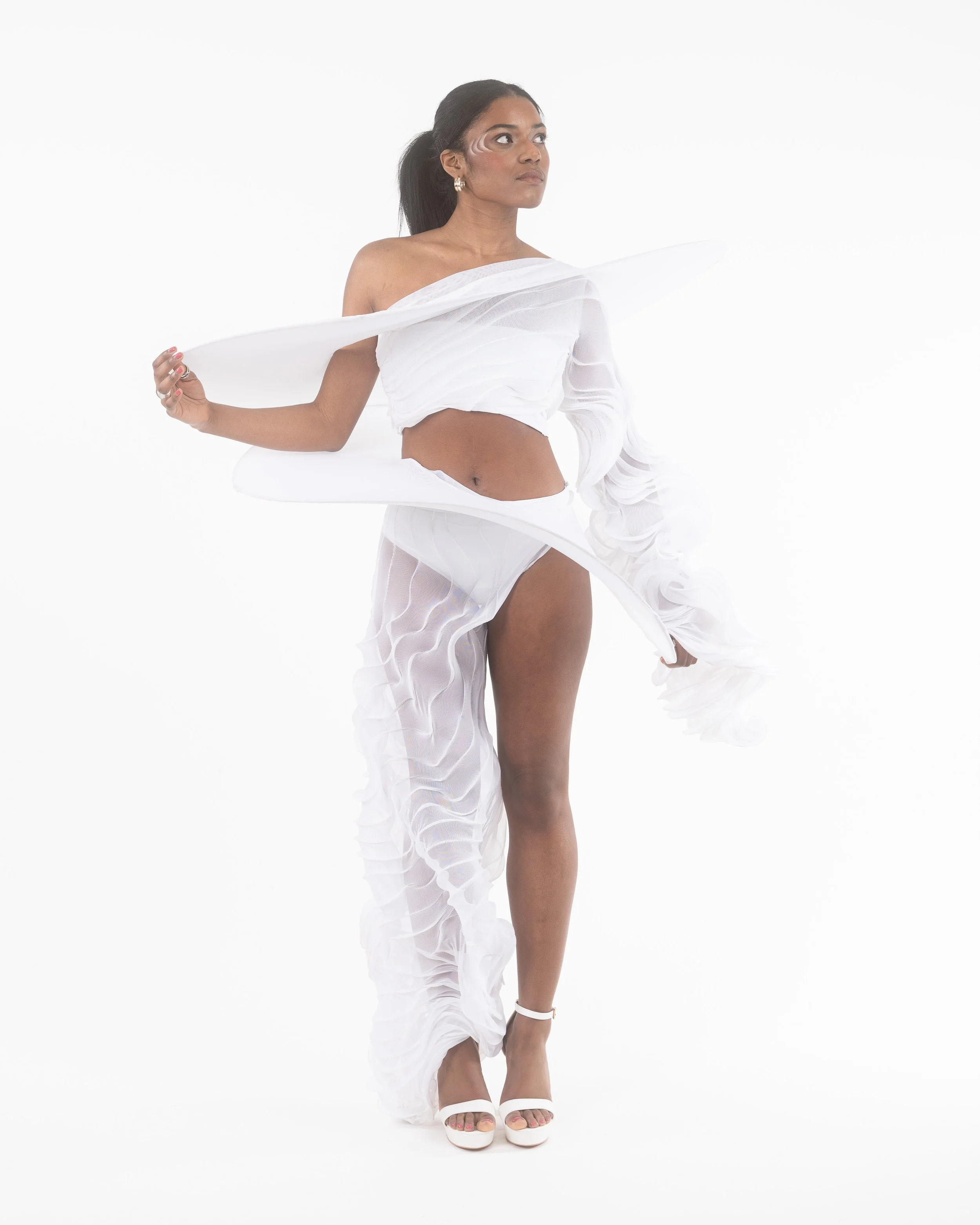 A woman wearing a white, semi-sheer, ruffled outfit and high heels standing against a white background.