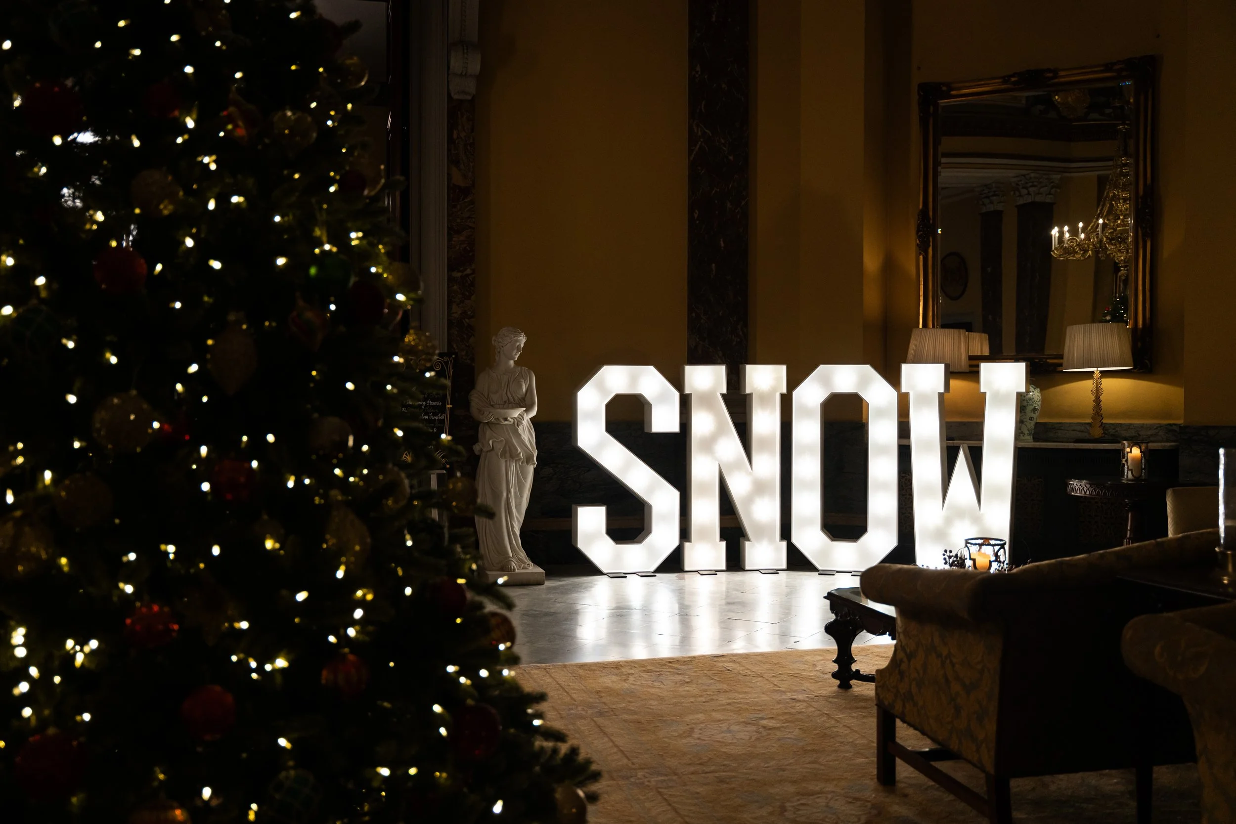 A decorated Christmas tree with lights on the left side of the image. In the background, there is a large illuminated sign spelling 'SNOW', a statue of a woman, and cozy lamps and furniture in a dimly lit room with elegant decor.