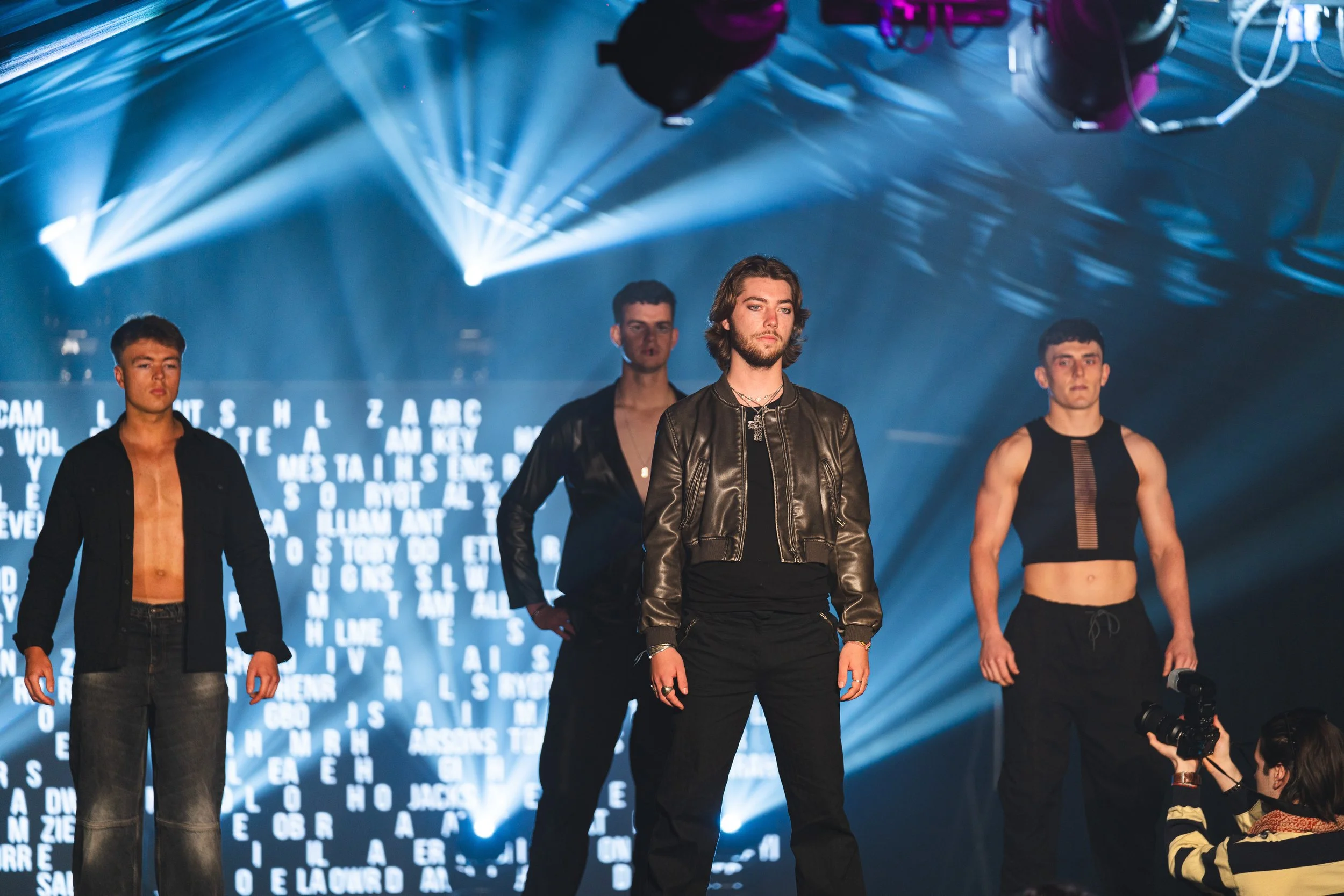 Four male models standing on a stage with digital text in the background, illuminated by stage lights, during a fashion show.