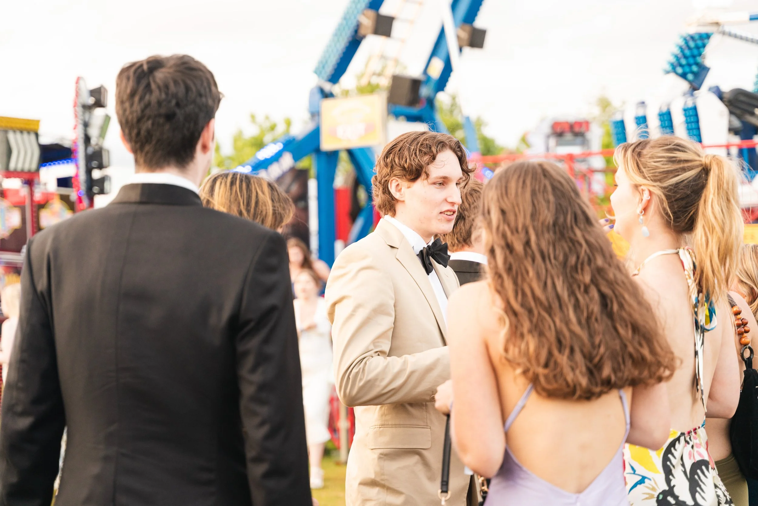 Group of young people in formal and casual attire talking at an outdoor fair with rides and booths in the background.
