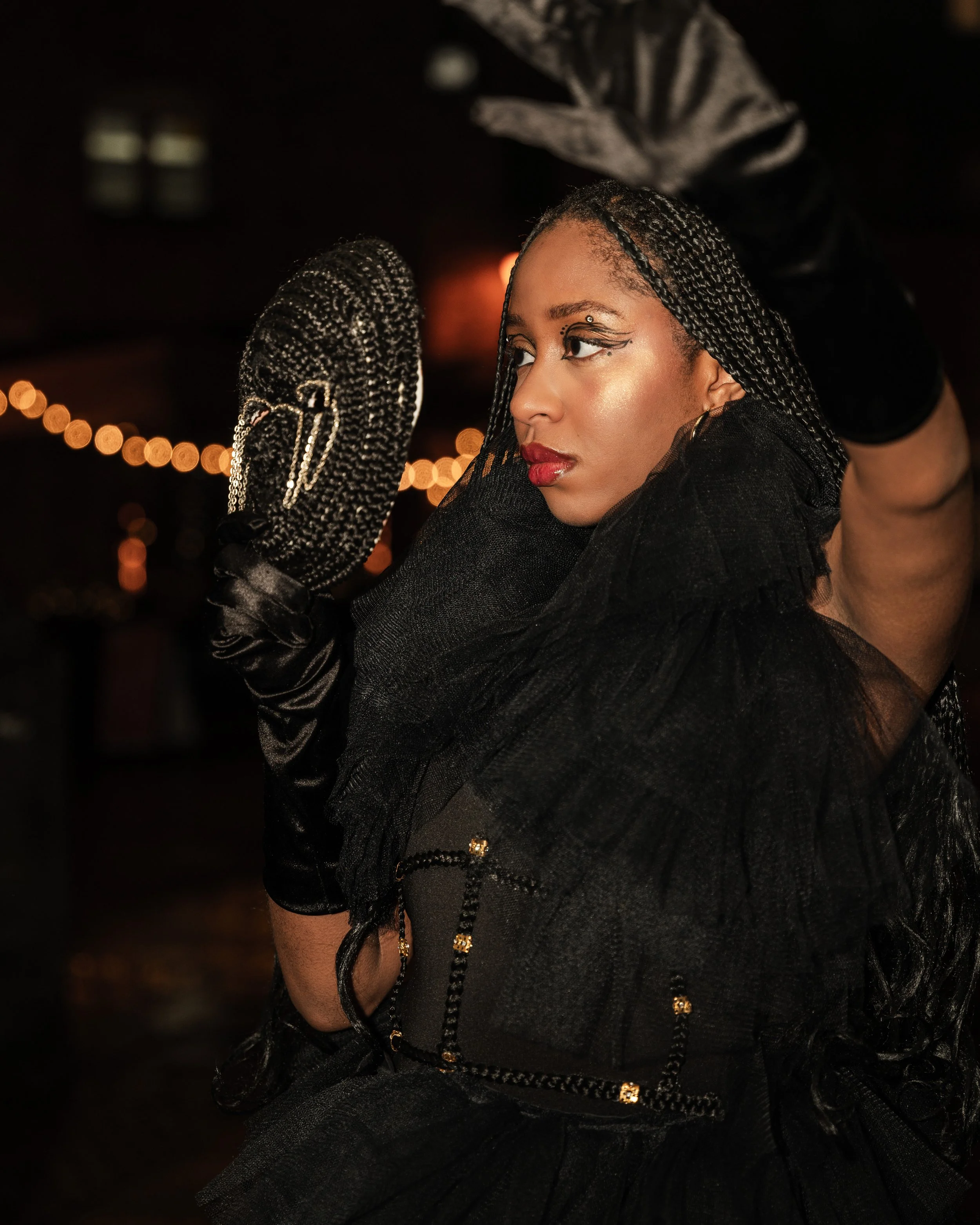 A woman with braided hair, bold makeup, and a black tulle dress is holding a decorative black clutch with a chain strap up to her face, and looking to the side with a serious expression in a dimly lit setting.