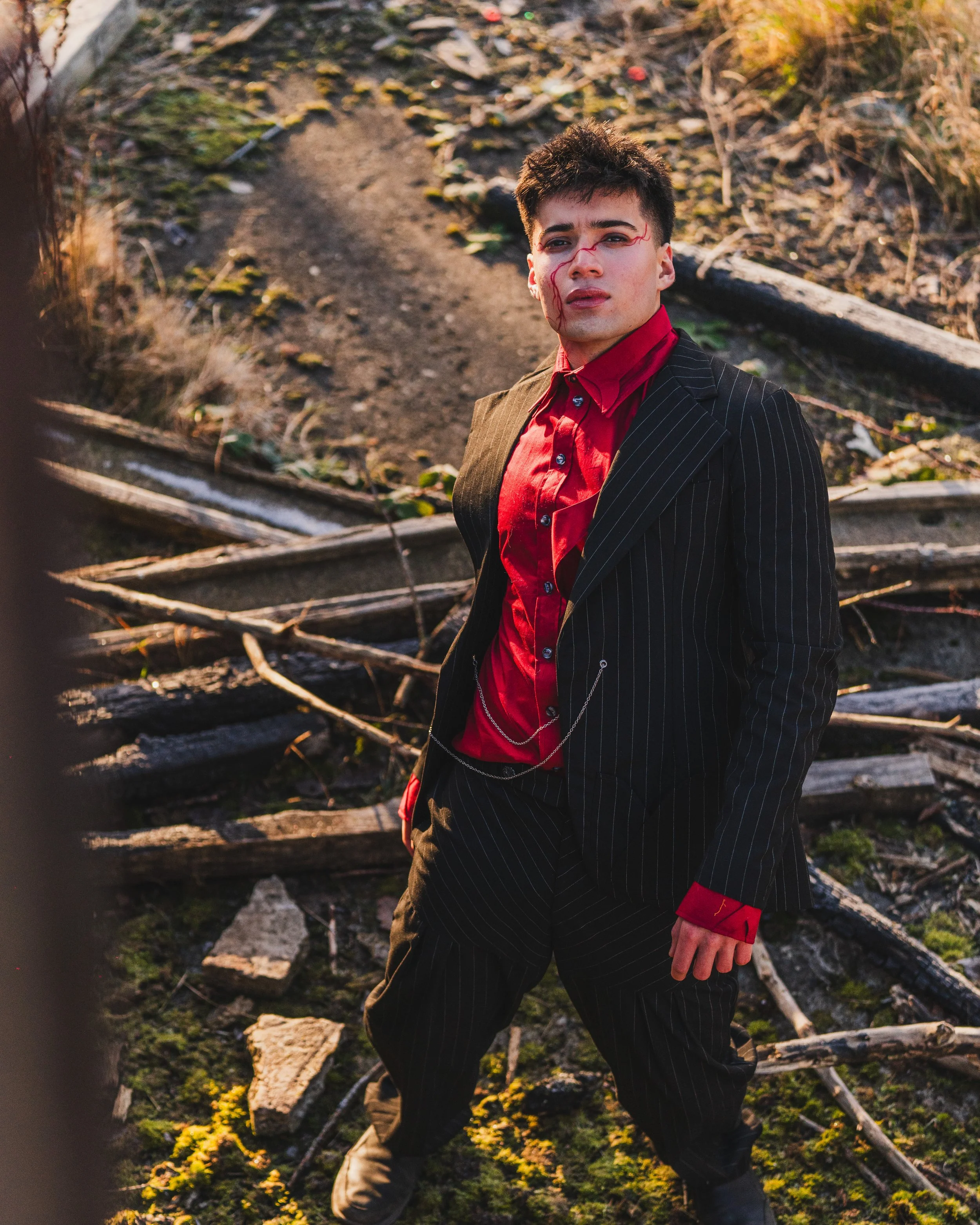 A young man dressed in a black pinstripe suit with a red shirt and red cuffs stands amidst fallen debris and broken wooden planks in a desolate outdoor setting, with facial makeup resembling blood streaks.