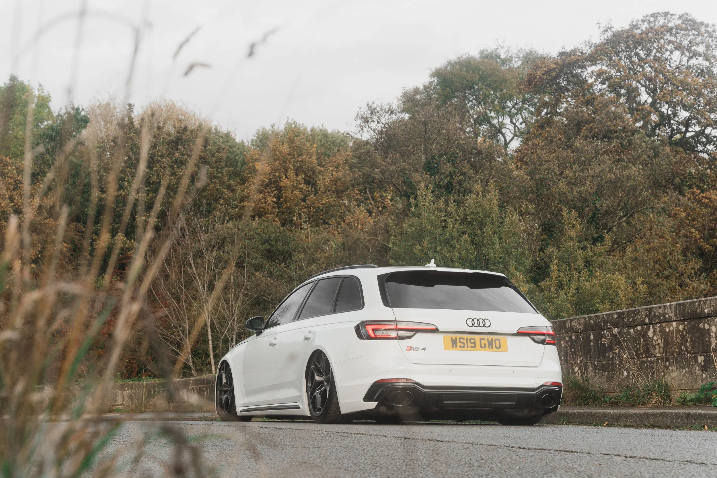 A white Audi RS 4 wagon parked on the side of a road with trees in the background and a concrete barrier on the right.