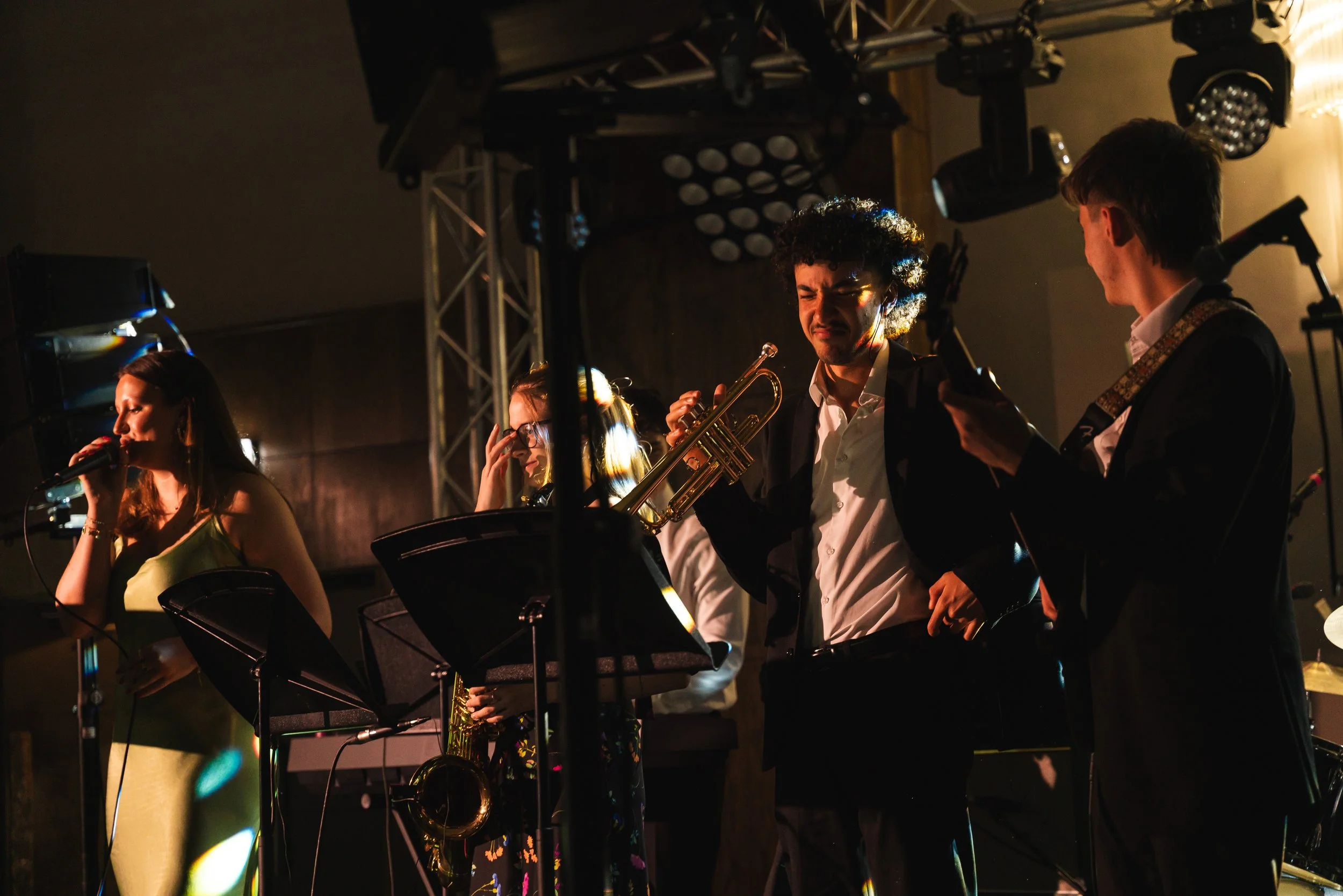 A live musical performance with five musicians on stage: a female singer, a female saxophonist, a male trumpet player, and two male guitarists, under stage lights.