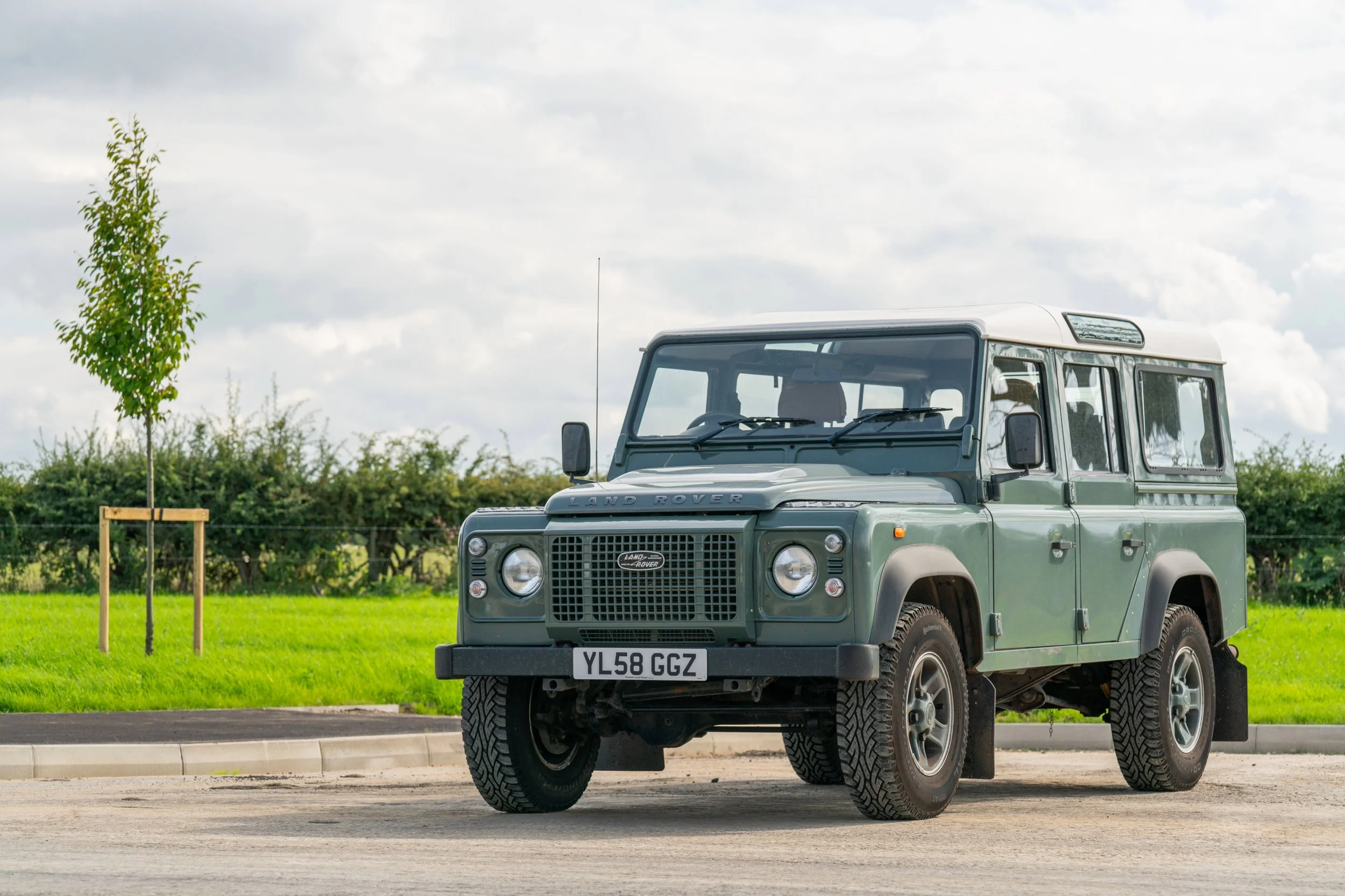 A classic green Land Rover Defender parked on a paved area with a grassy field, trees, and cloudy sky in the background.