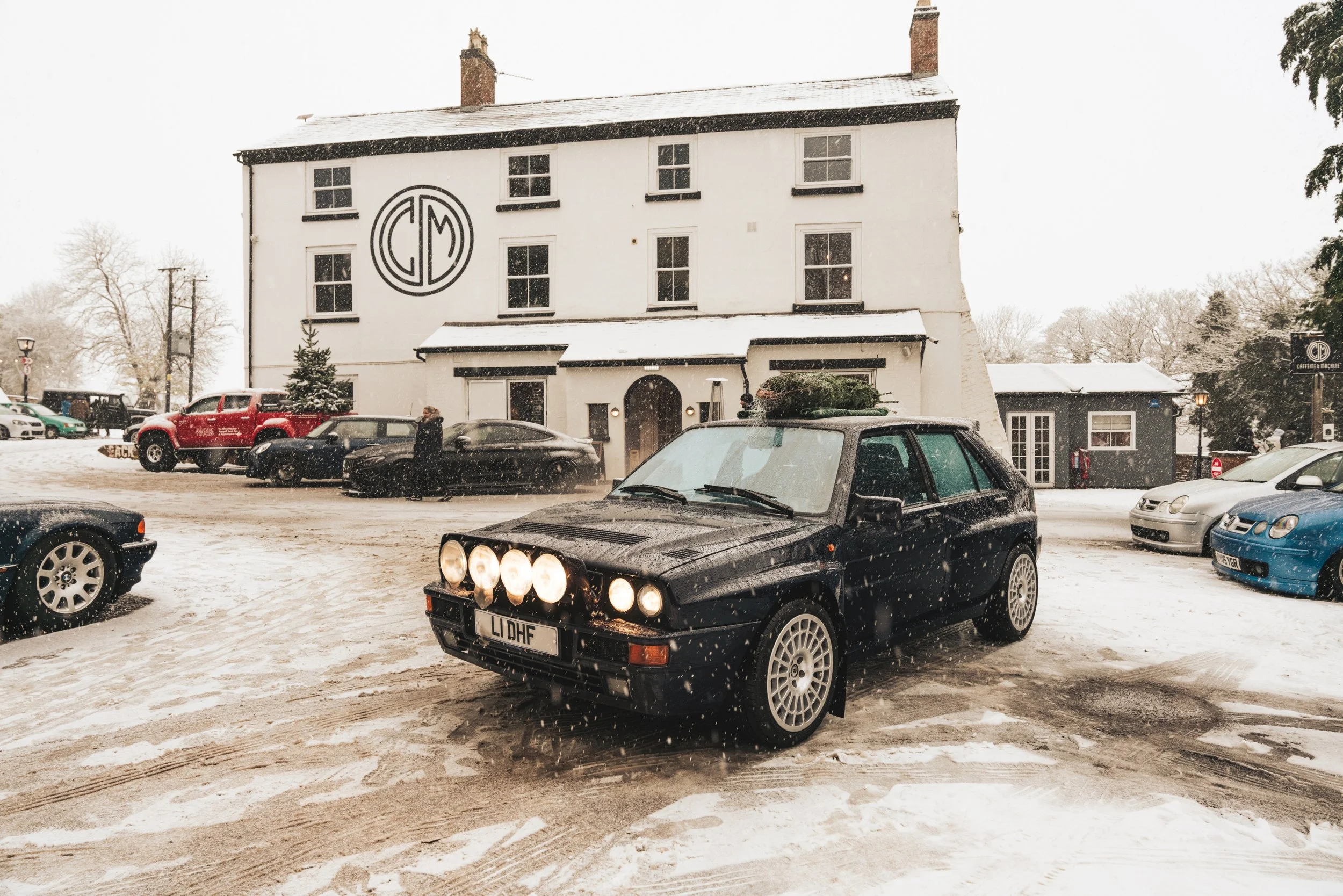A black vintage car with four large front lights parked on a snowy street in front of a white building with a logo on it, with other cars and trees in the background.