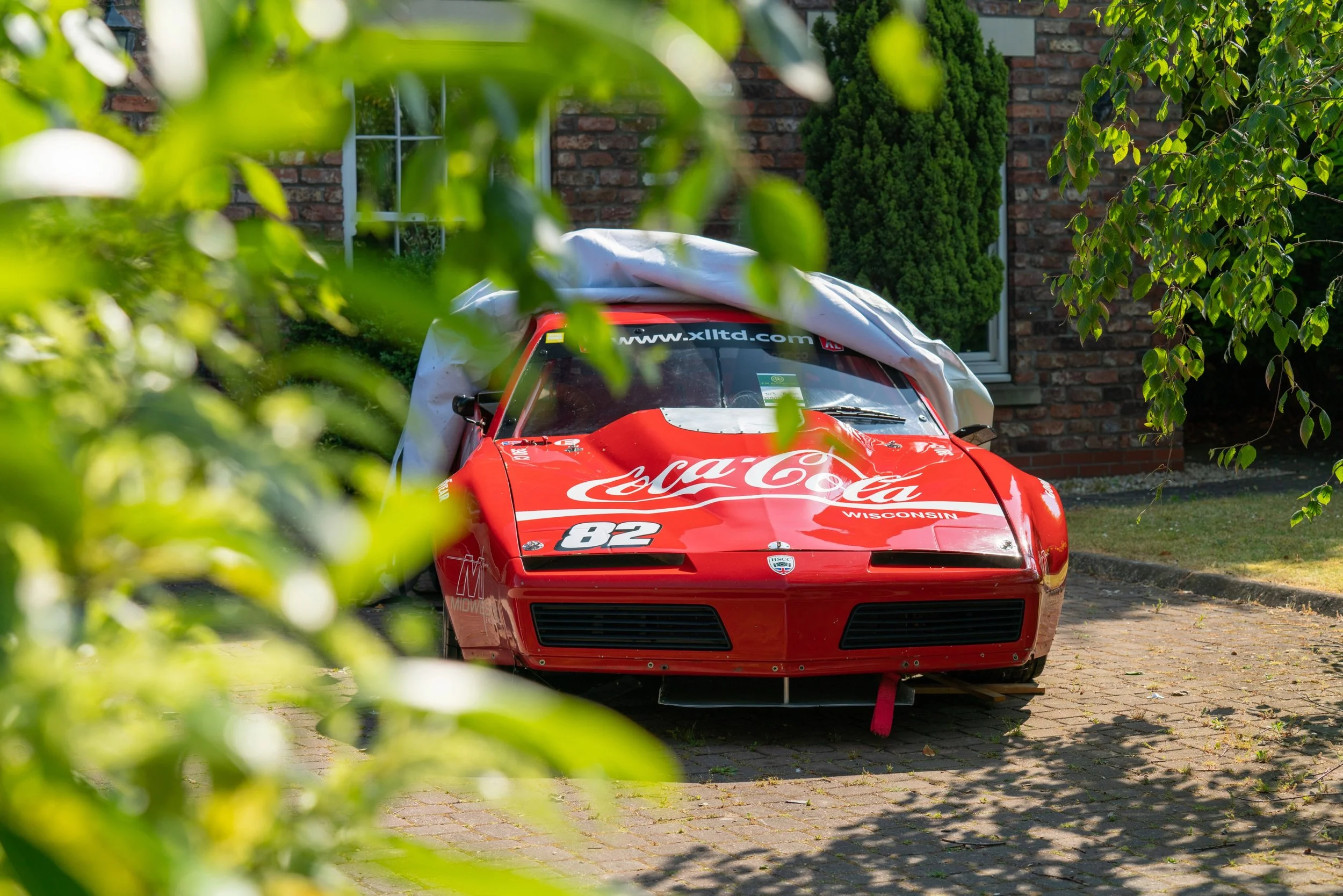 Red racing car with Coca-Cola branding and the number 82, parked on a brick driveway, partially obscured by green foliage and in front of a brick house.