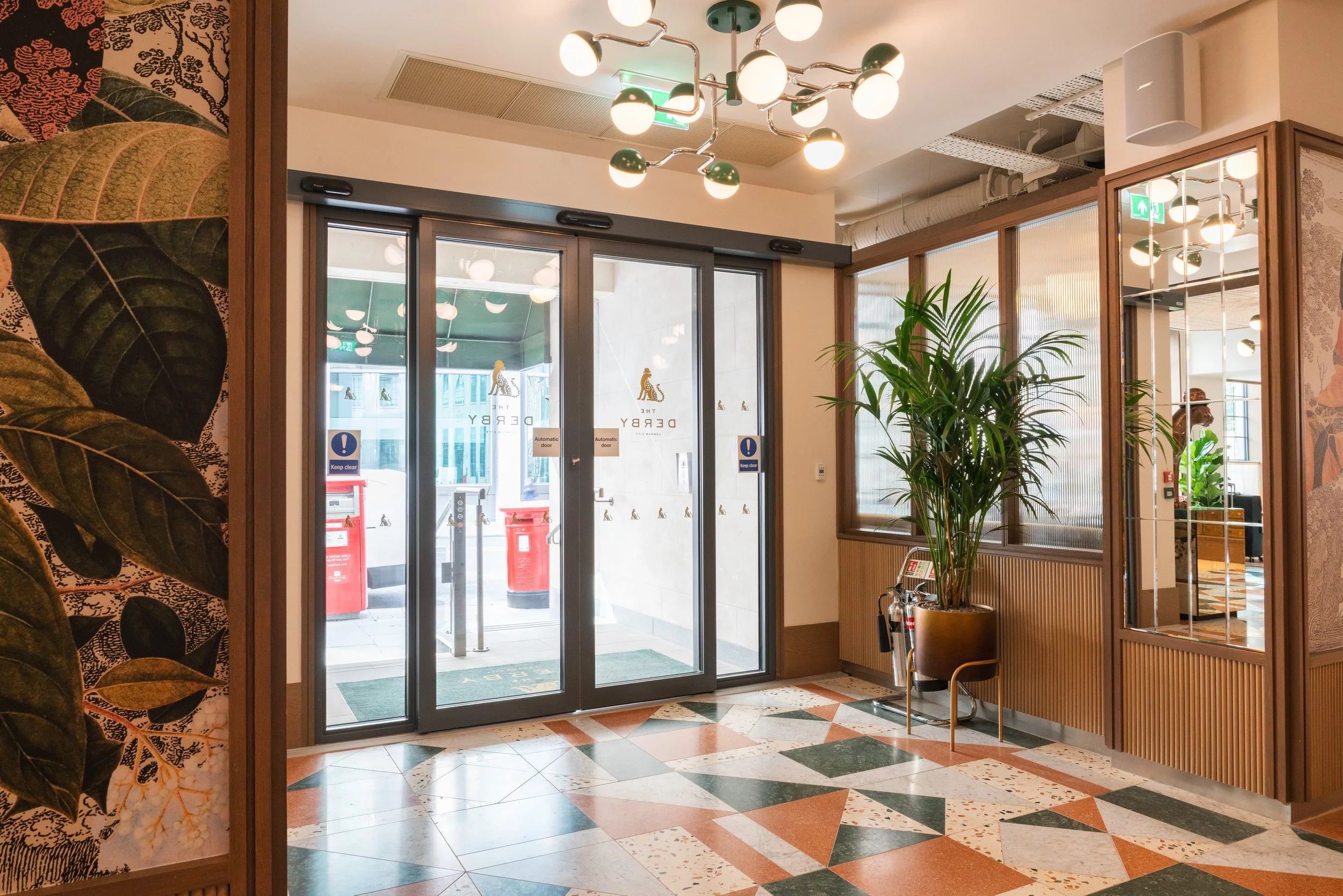 Indoor space with glass doors labeled 'The Derby' and decorative plants, with patterned tile flooring and stylish lighting fixtures.