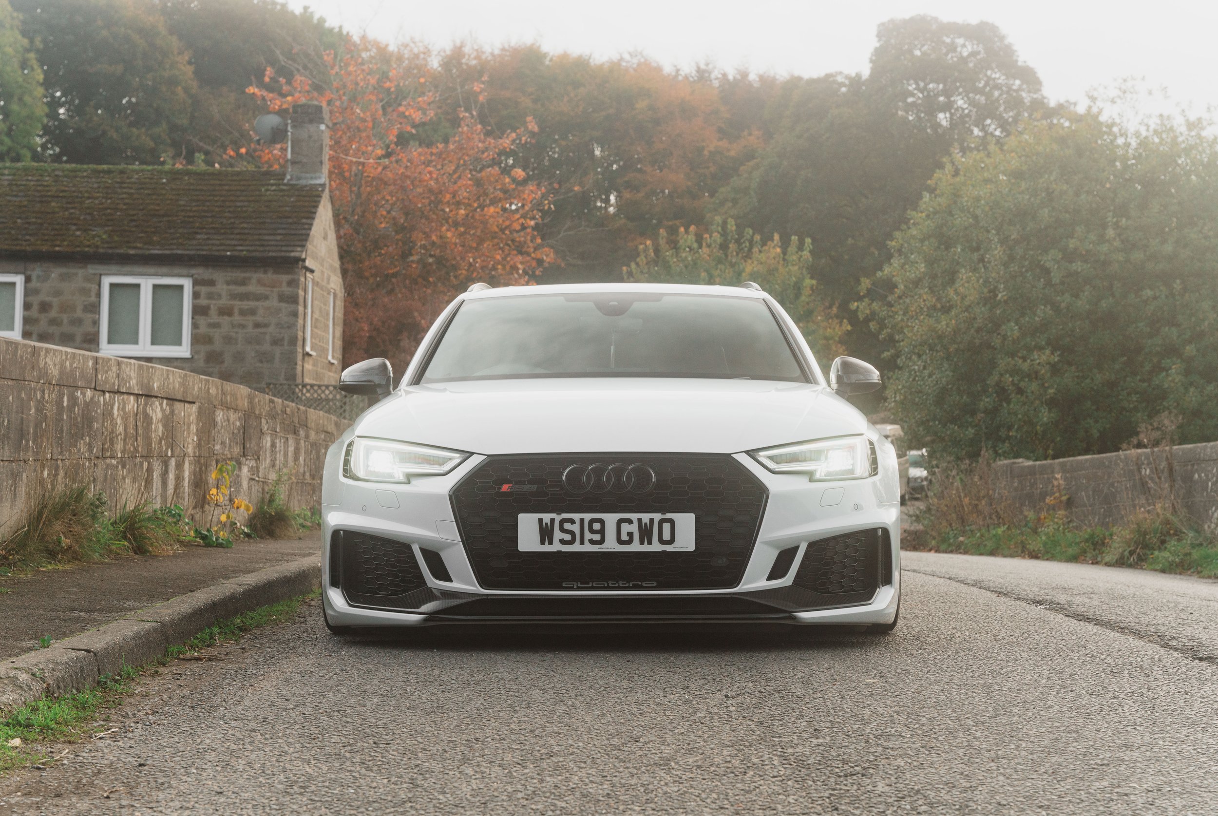 Front view of a white Audi Quattro car parked on a narrow residential street with autumn trees and houses in the background.