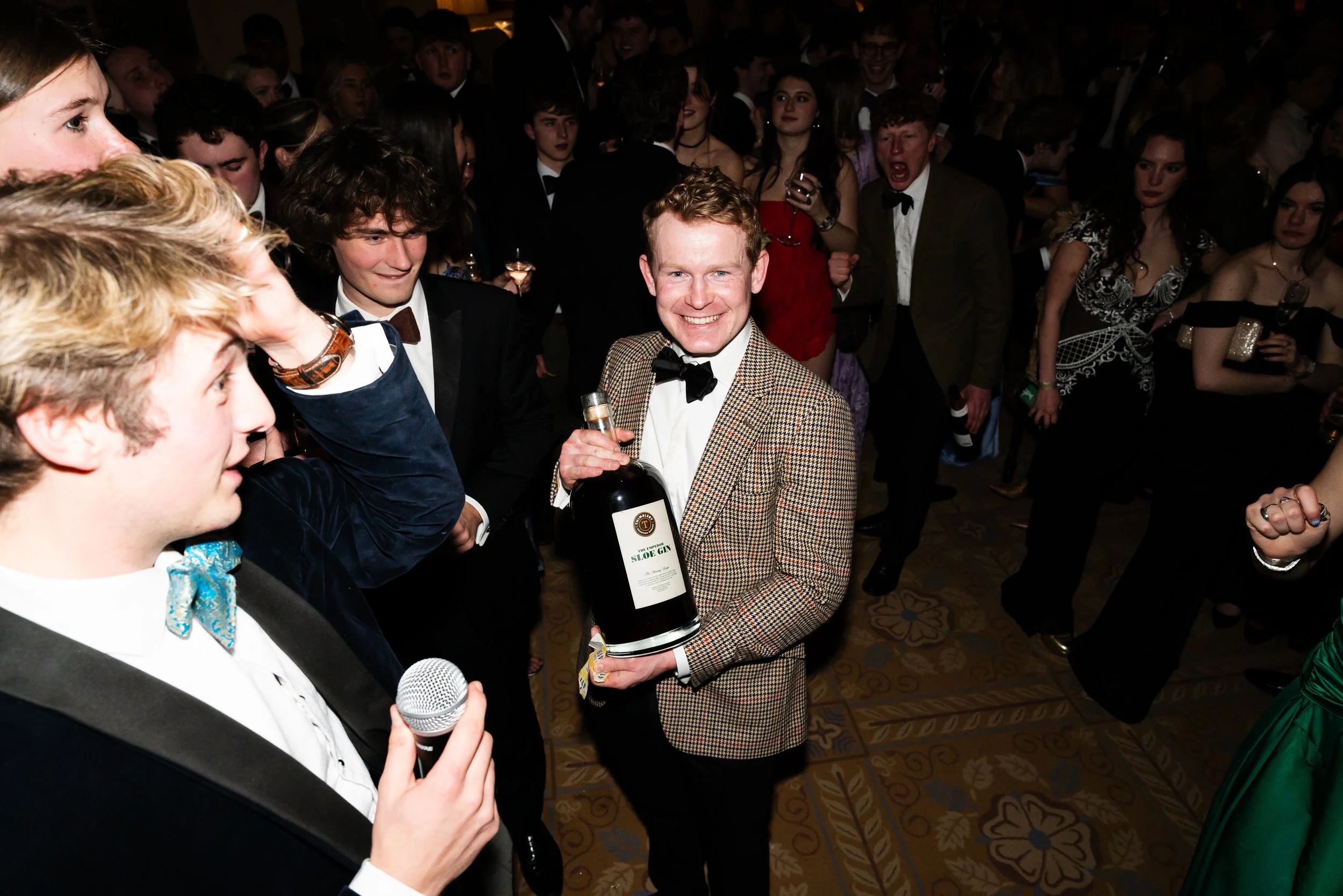 A young man in a checkered blazer and black bow tie is smiling and holding a large bottle of gin at a party, surrounded by people in formal attire.