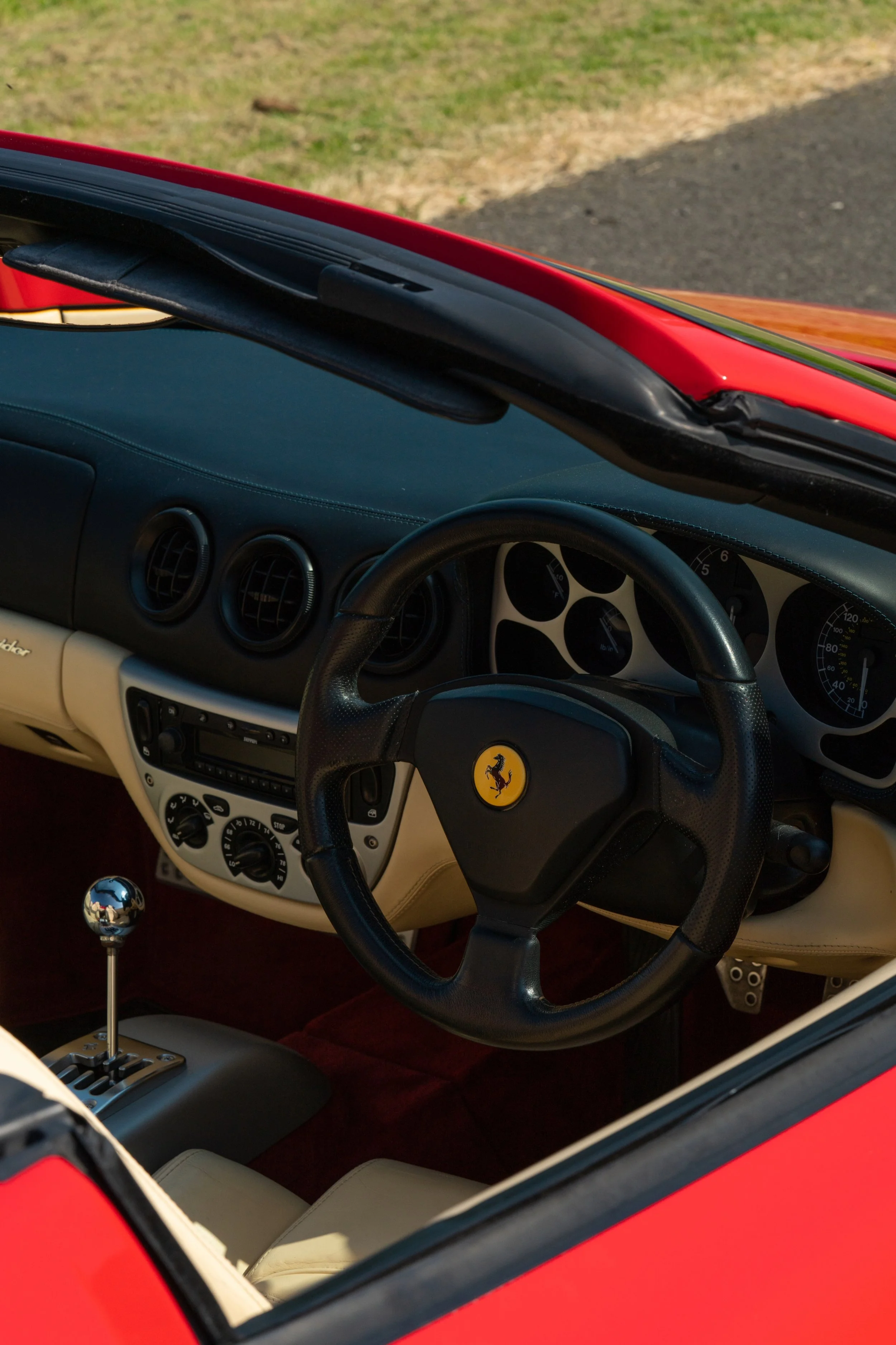 Interior of a red Ferrari sports car, showing the steering wheel with a Ferrari logo, dashboard, gear shift, and cream-colored seats.