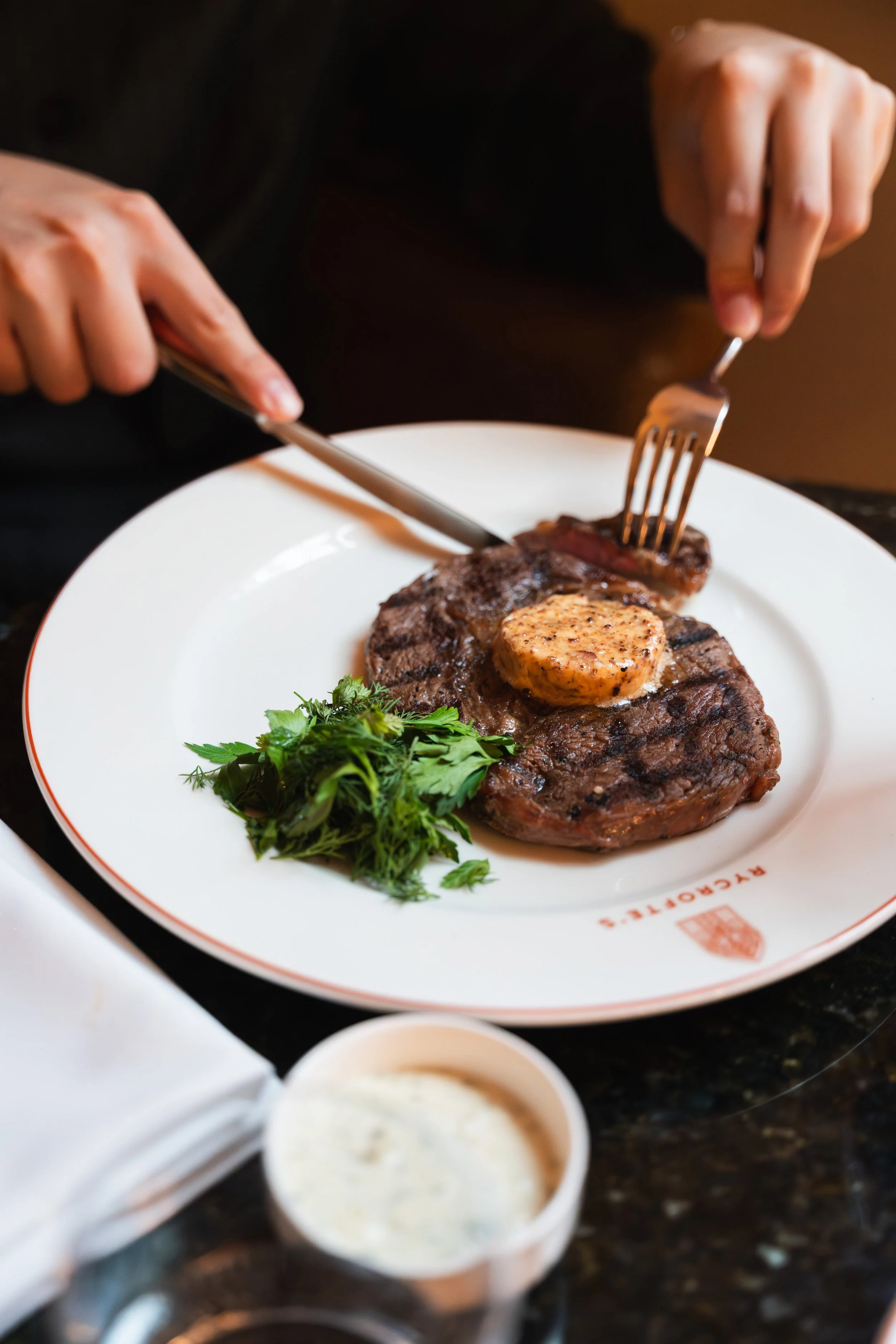 A plate with a grilled steak topped with a small piece of butter and garnished with fresh herbs, with small cups of dipping sauces in the foreground.