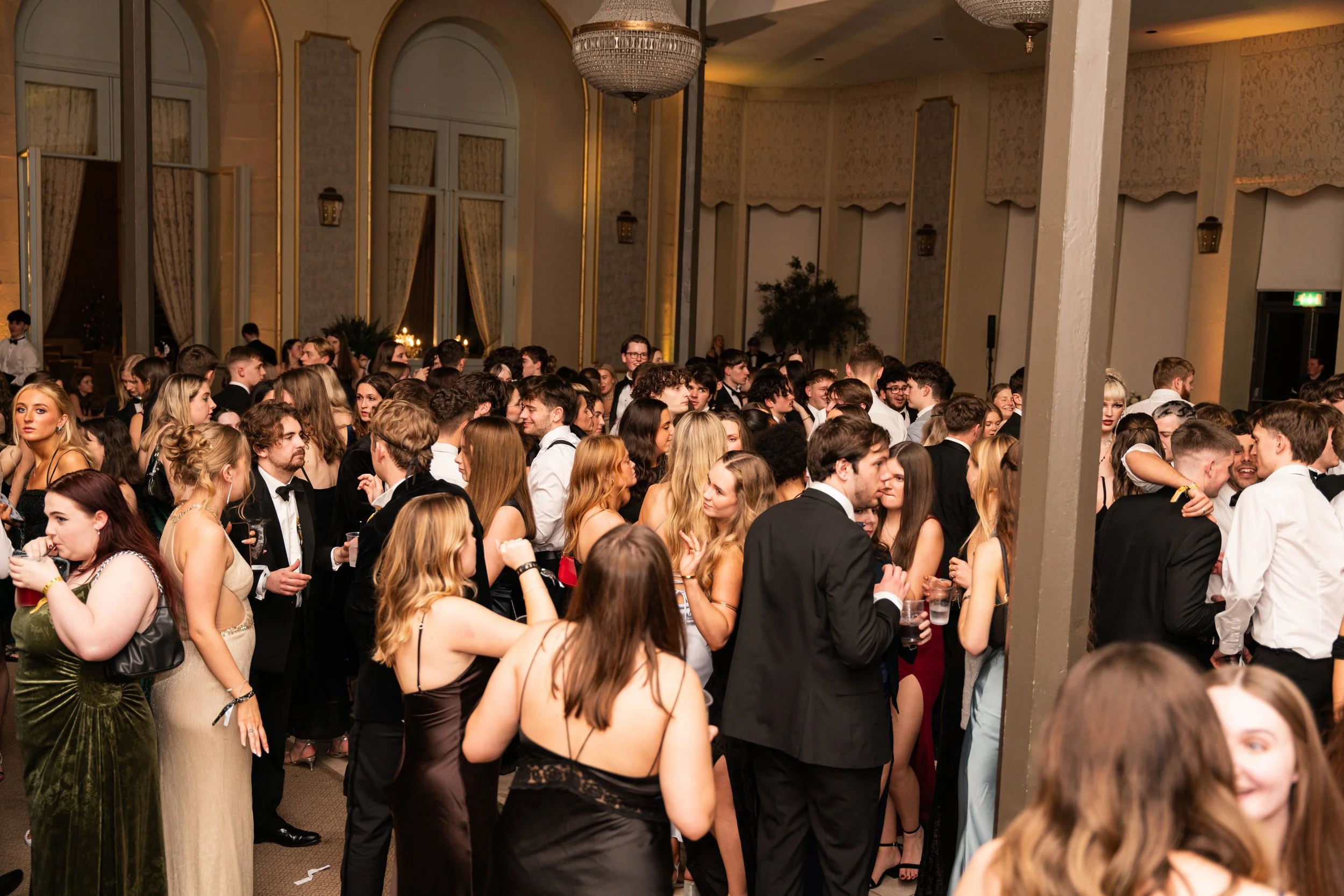 Crowd of people dressed in formal attire at a large indoor event, dancing and socializing in a ballroom with ornate decorations and high ceilings.