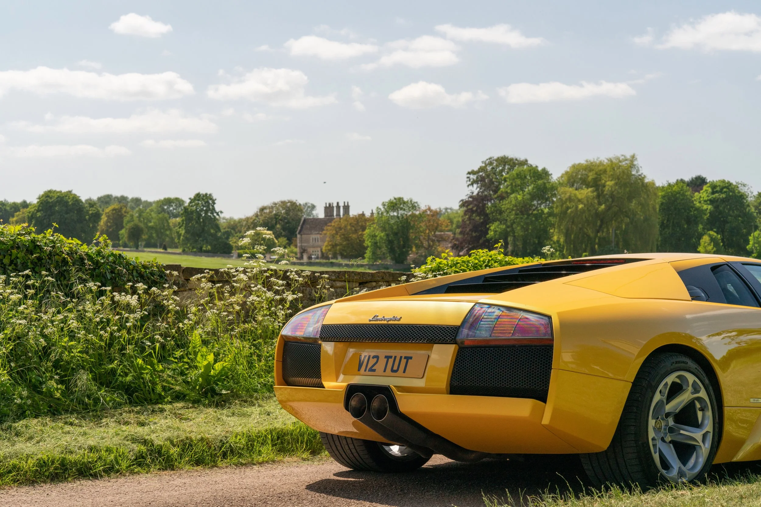 Yellow Lamborghini sports car parked on a countryside road with green trees and a stone house in the background under a partly cloudy sky.