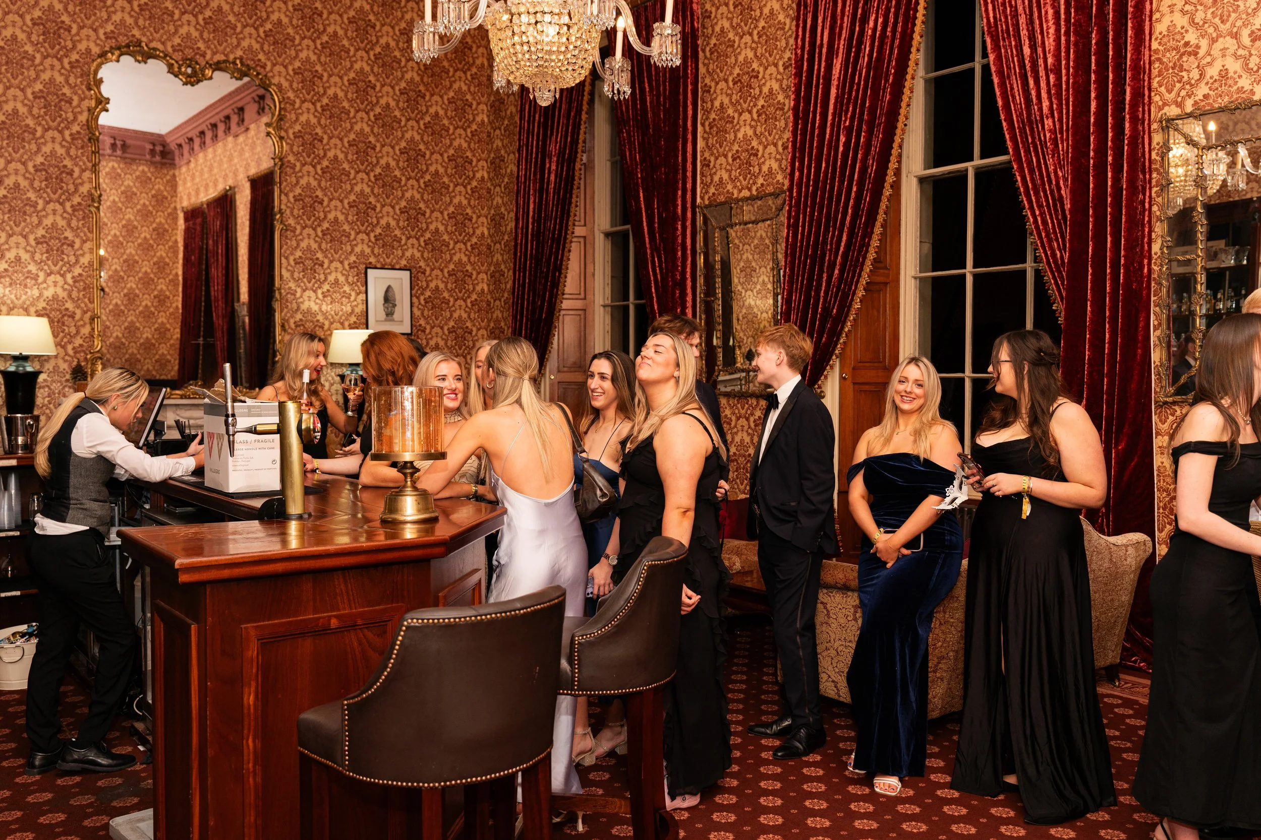 Elegant party scene with women in evening gowns in line at a hotel bar, with a bartender preparing drinks, ornate wallpaper, large red curtains, and a chandelier in a luxurious room.