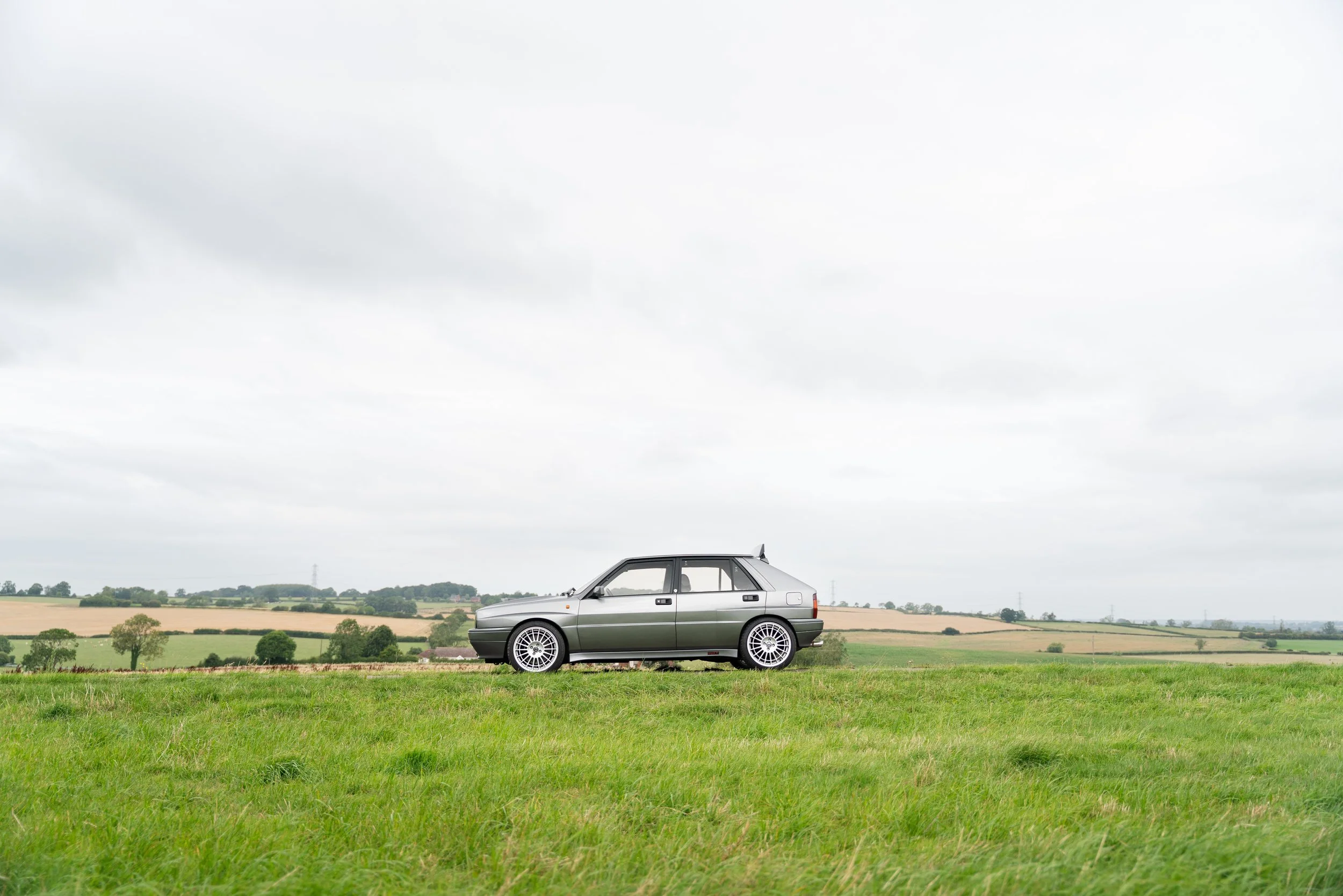A silver hatchback car parked on a grassy field with a rural landscape and cloudy sky in the background.