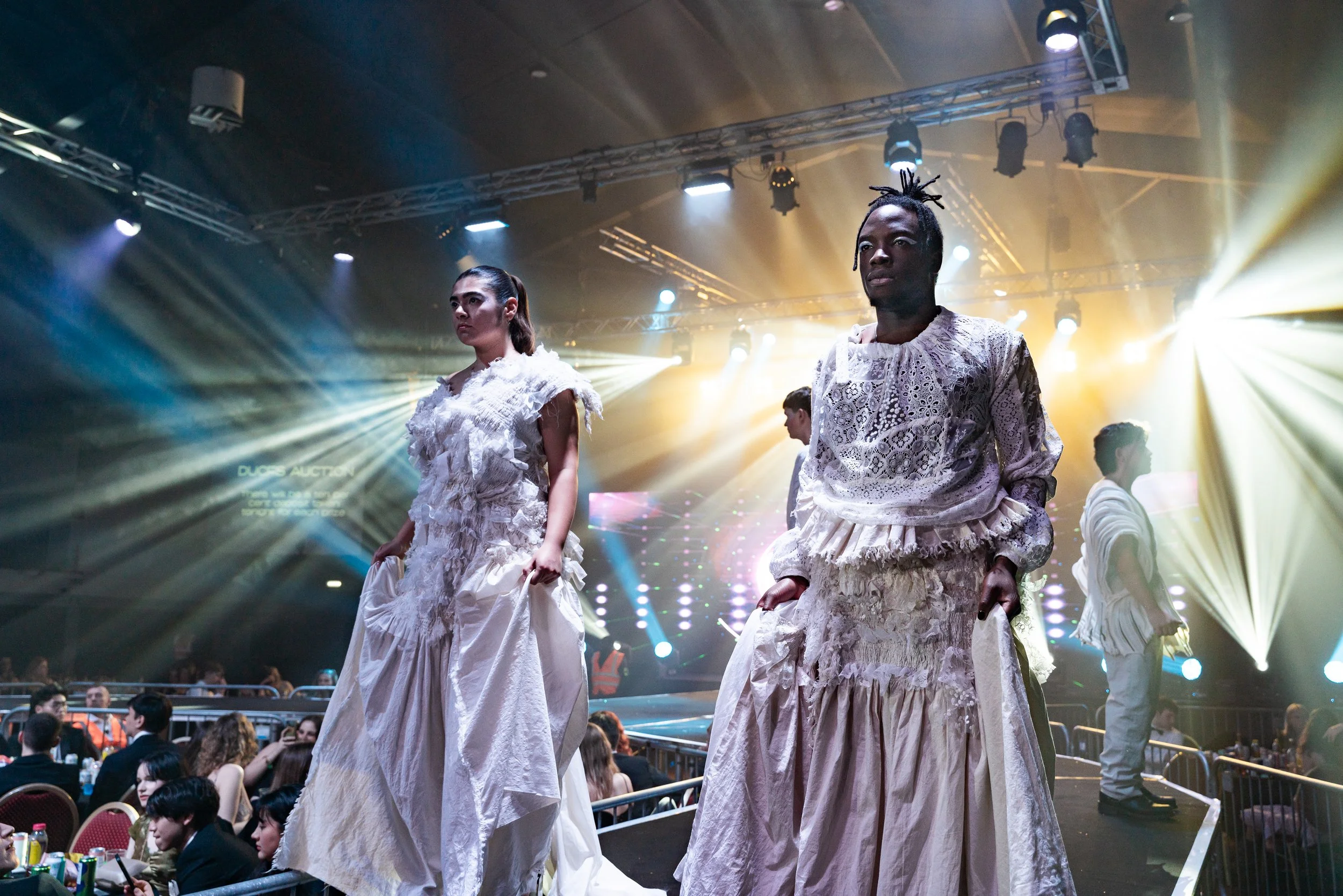 Models walking on a runway at a fashion show, with bright stage lights and audience watching.