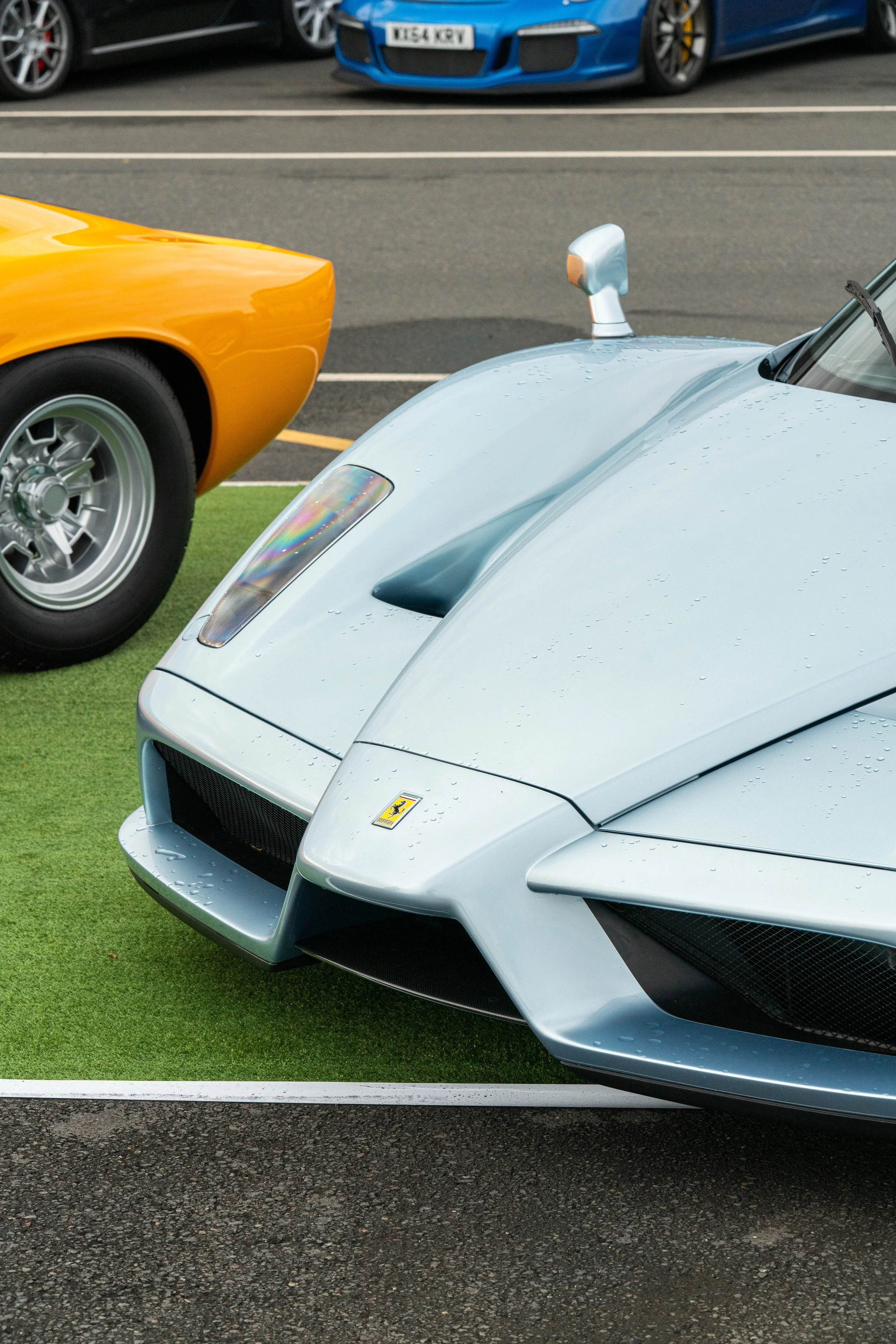 Close-up of a silver Ferrari sports car with water droplets on its surface, parked on a green patch, with a yellow car nearby and other cars in the background.