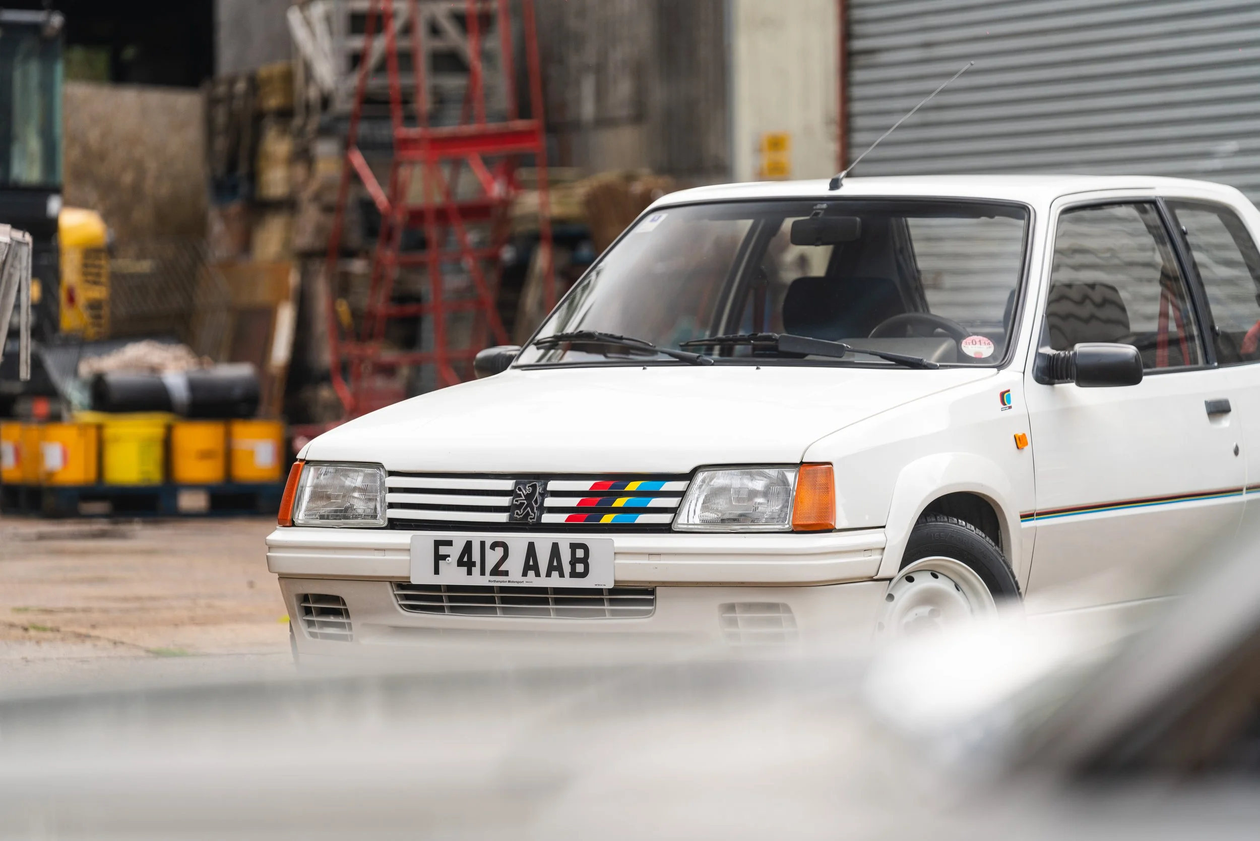 A white vintage Peugeot car with a black grille, colorful stripes, and UK license plate parked in an industrial yard with storage shelves and equipment in the background.