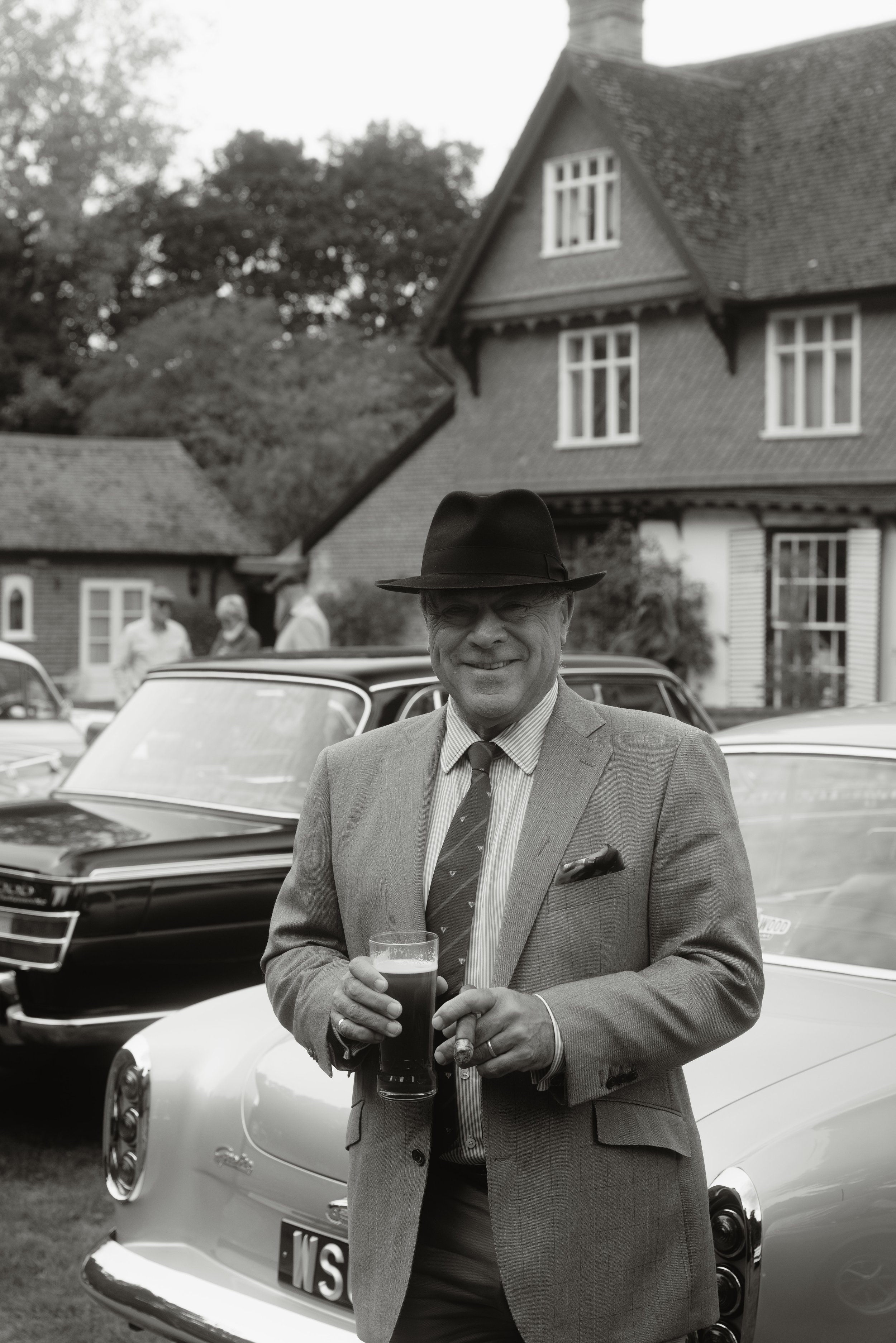 Man in a suit and fedora hat smiling while holding a pint glass and cigar, standing in front of vintage cars at an outdoor gathering
