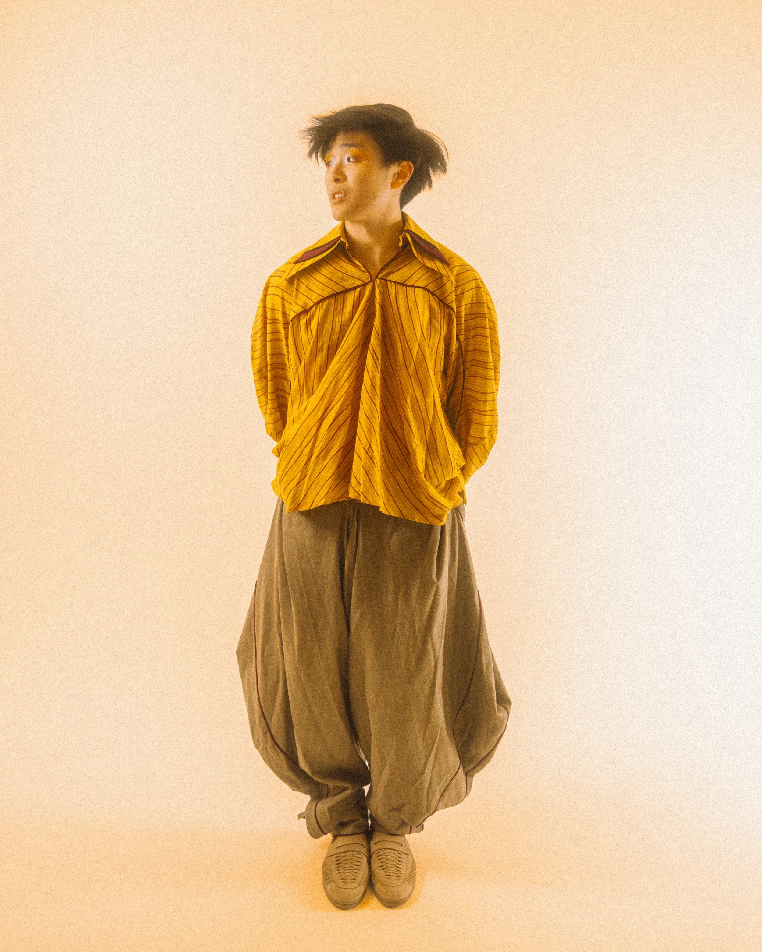 Person with messy hair wearing a yellow striped oversized shirt, baggy gray pants, and sneakers, standing against a plain light-colored background.