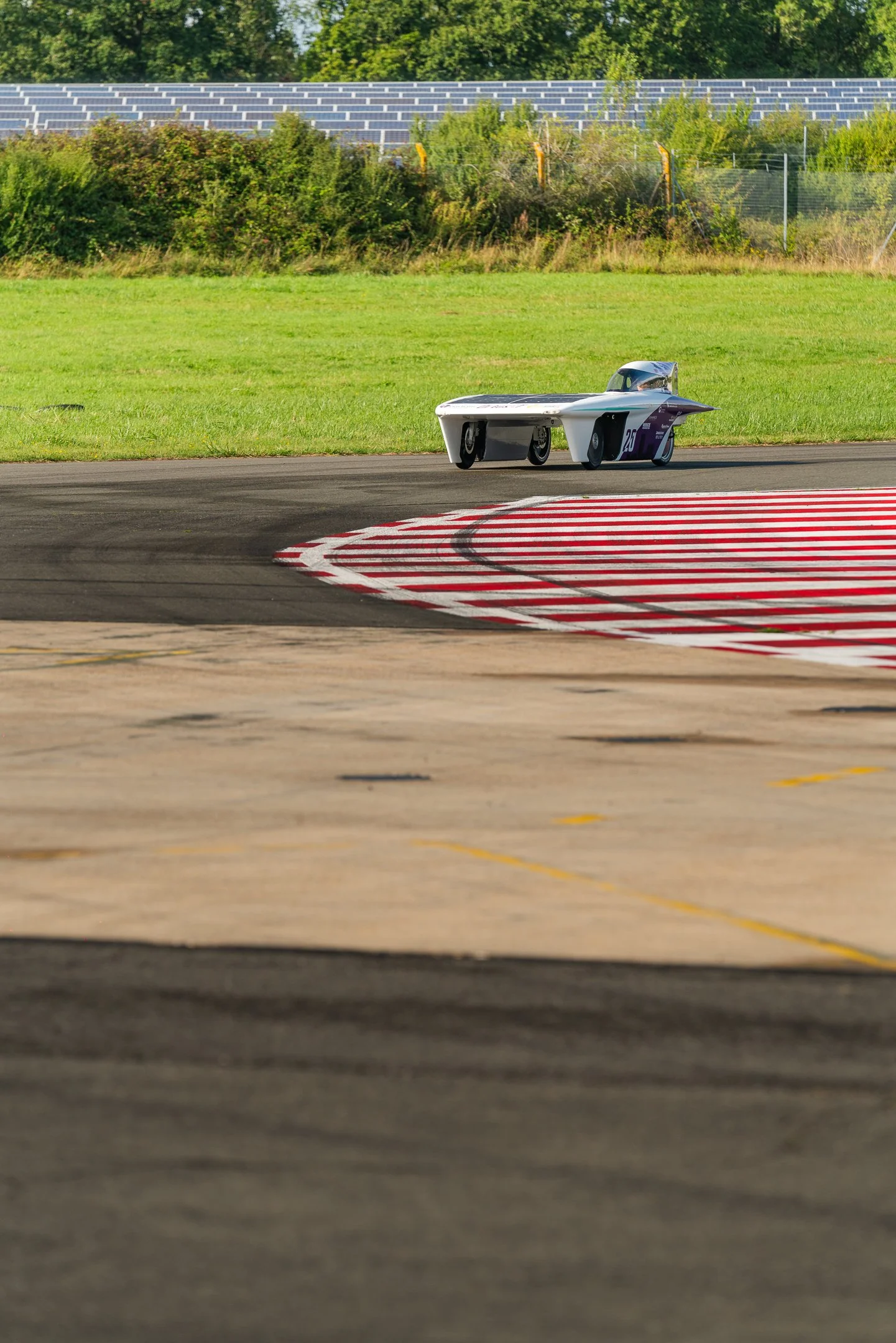 A small, vintage race car on a racetrack with red and white curbing, green grass, bushes, and solar panels in the background.