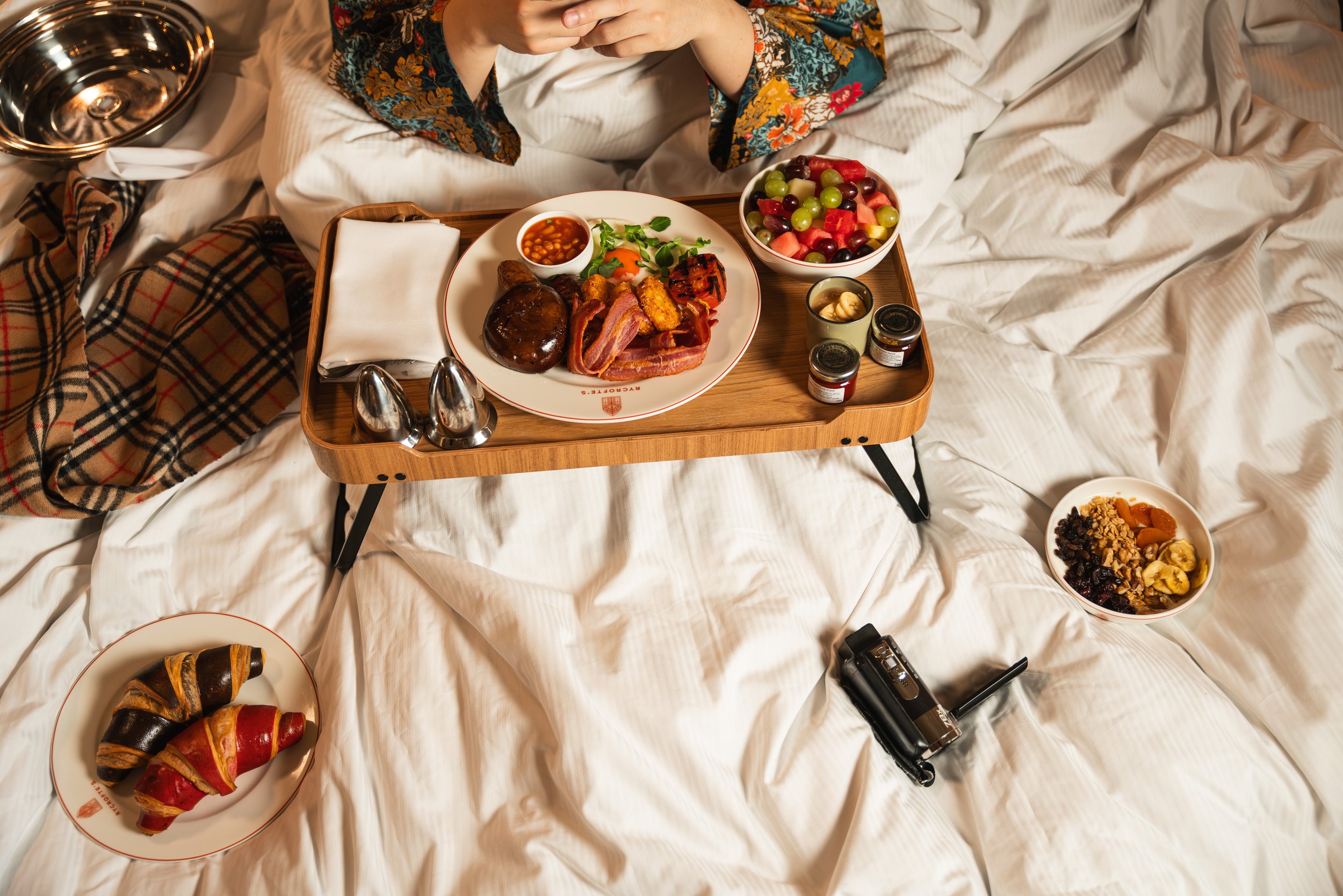 A tray with breakfast foods including baked beans, grilled tomatoes, bacon, mushrooms, and a side salad, on a bed with a person in colorful pajamas in the background. There are bowls of fruit and granola, and croissants on separate plates, with a cam